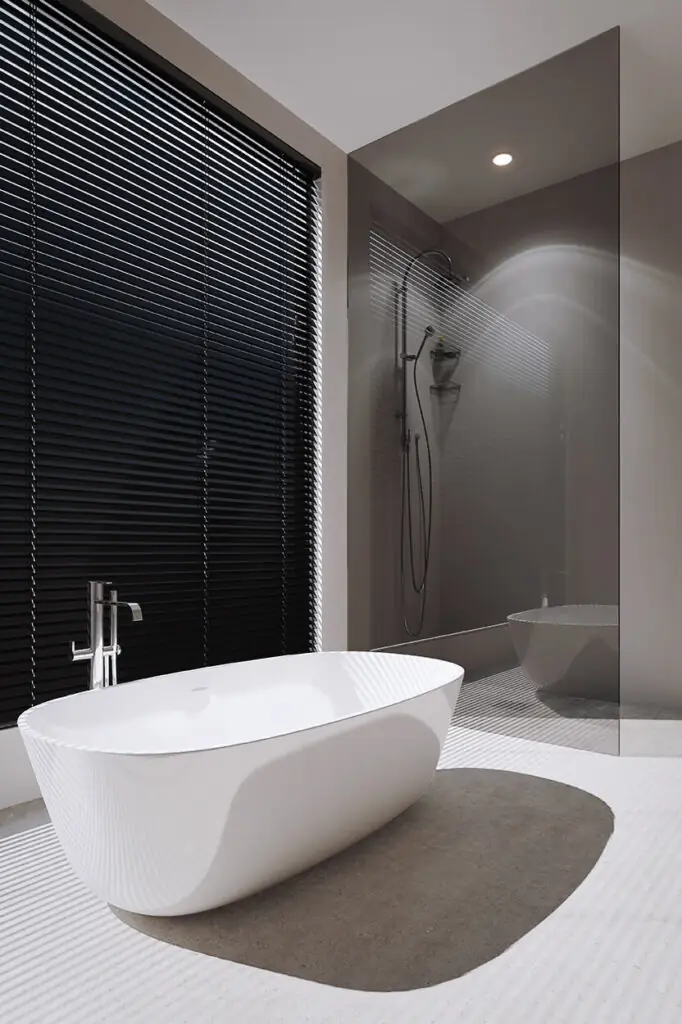 Freestanding bathtub beside a window with black horizontal blinds casting striped shadows