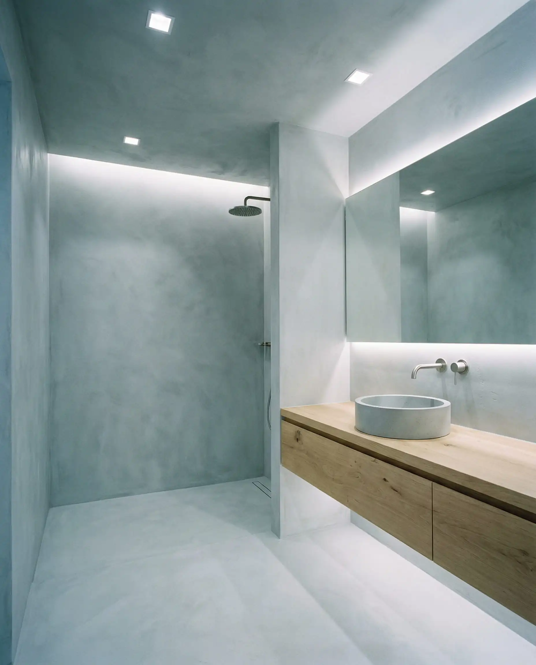 A modern, spa-like bathroom featuring a continuous, joint-free micro-cement (Beton Cire) coating on all walls and the floor in a pale, dusty cool grey. This bathroom features Pale cool grey, light oak wood, and brushed silver fixtures tones and a sculpted, serene, modern european design atmosphere.