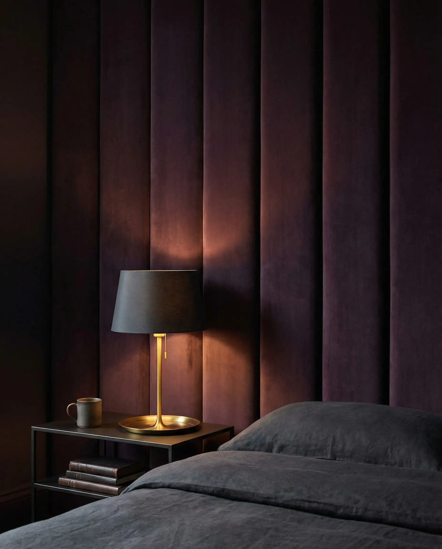 A dramatic bedroom with floor-to-ceiling upholstered fabric panels in a deep 'Divine Damson' (blackened plum/purple) matte velvet, replacing a traditional headboard. This bedroom features Deep matte plum, brushed gold brass, and dark charcoal grey tones and a introspective, hotel vibe, cocooning atmosphere.