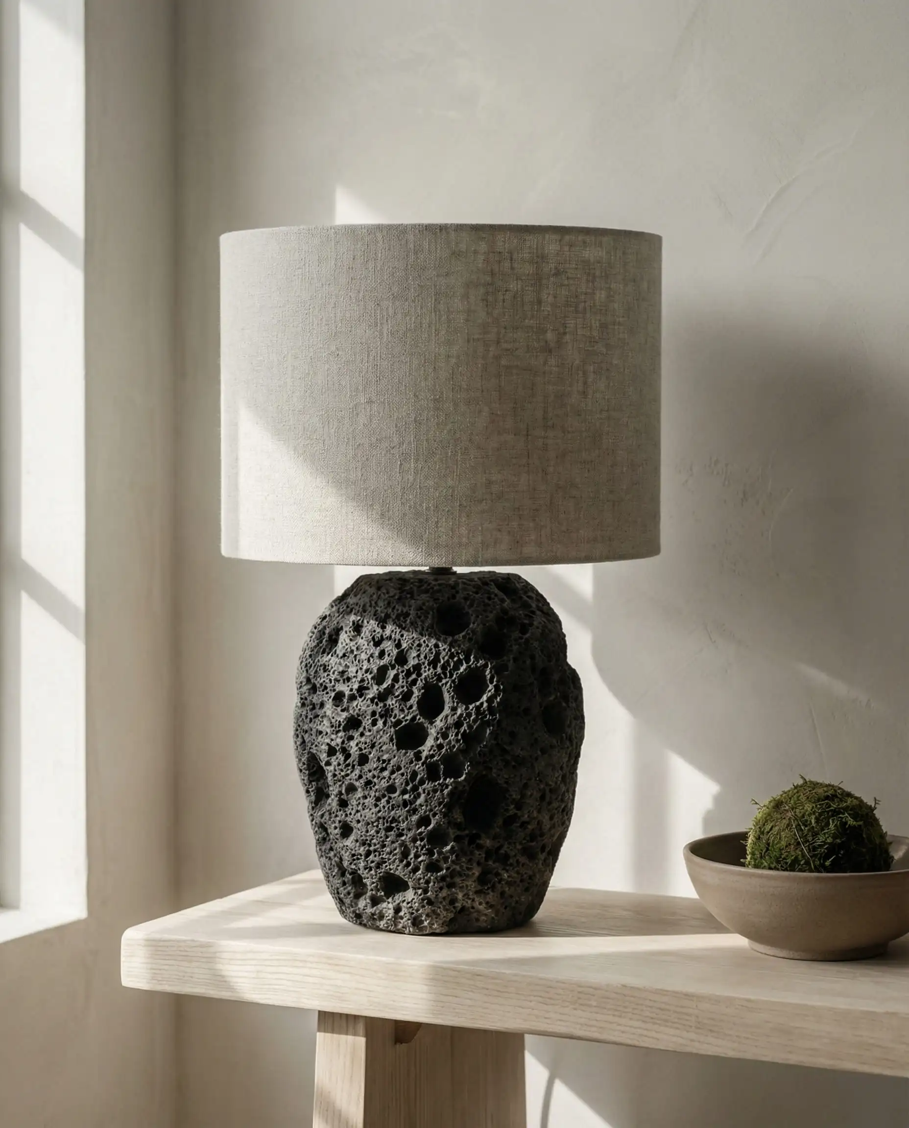 Porous black lava stone table lamp with a raw linen shade, showcasing wabi-sabi texture in a sunlit room.