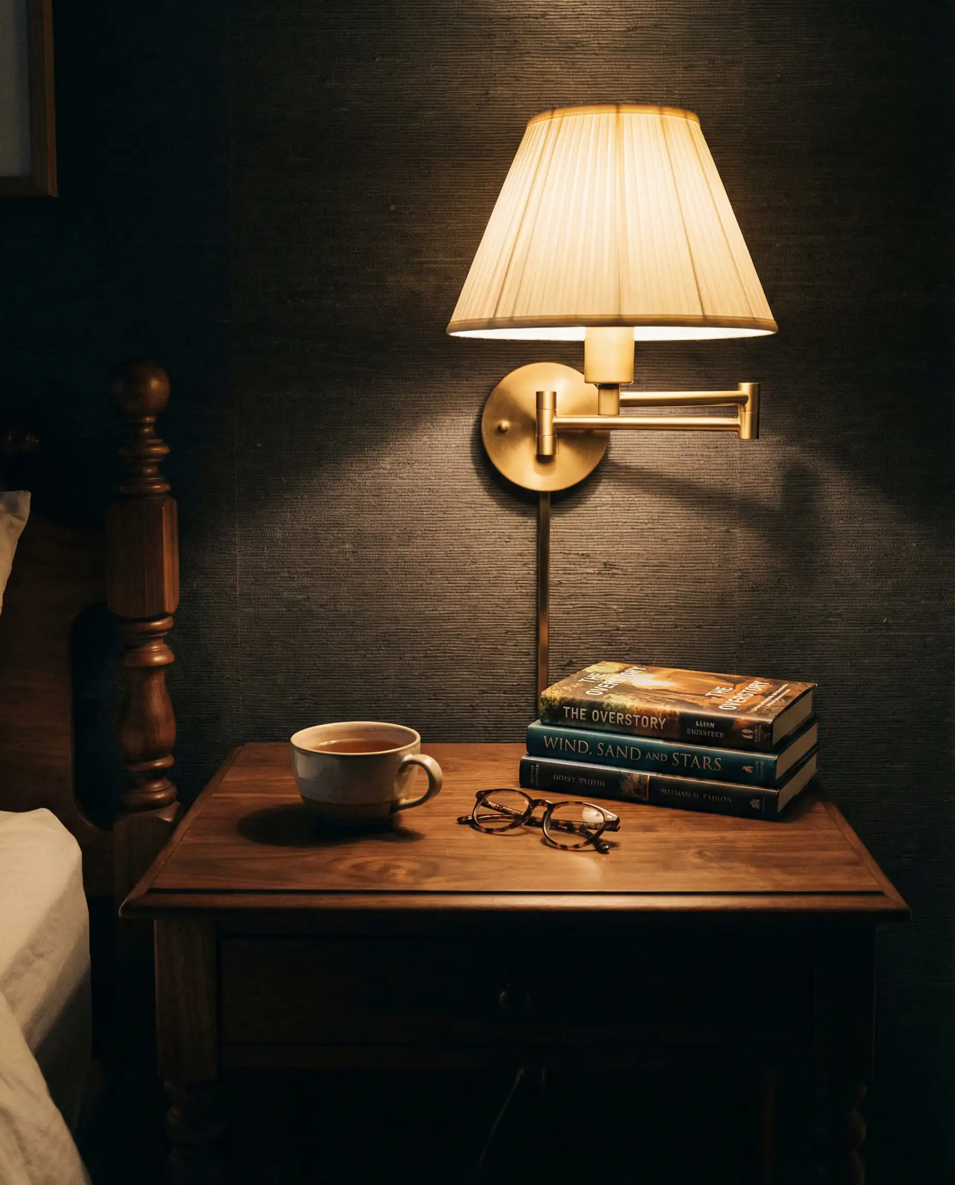 Bedside detail showing a walnut nightstand lit by a brass wall sconce with a pleated shade.
