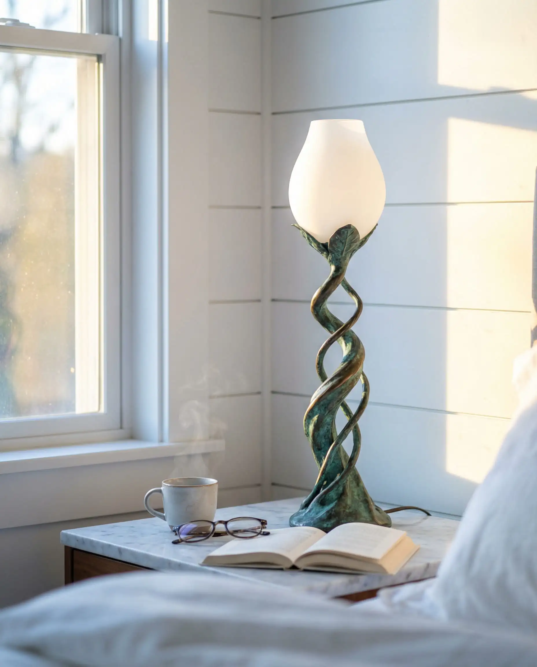 Whimsical bronze table lamp designed to look like a sprouting plant stem holding a frosted glass bud, styled on a marble bedside table.