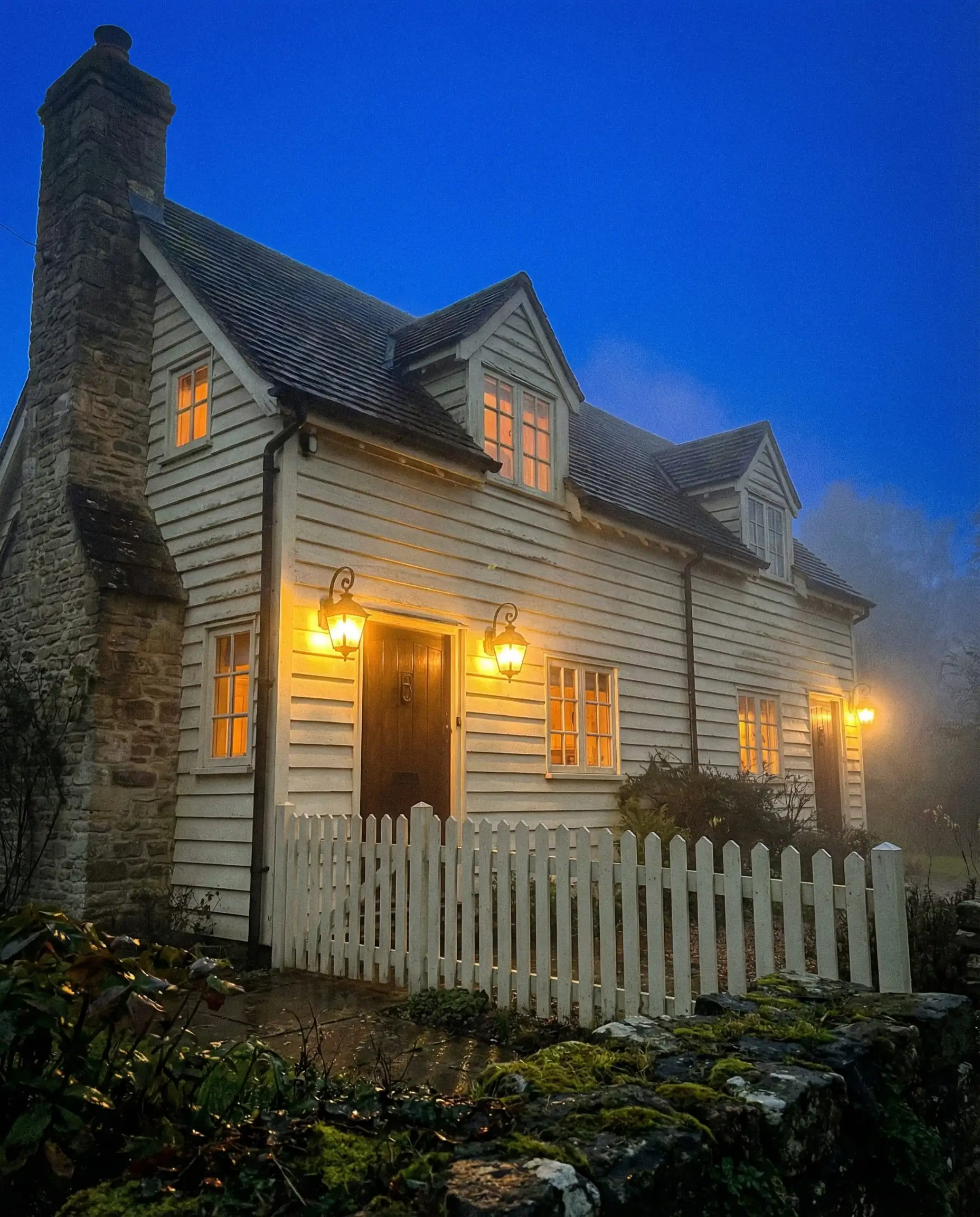 A cozy English cottage painted in warm off-white, illuminated by exterior sconces at sunset showing the paint's warm undertones.