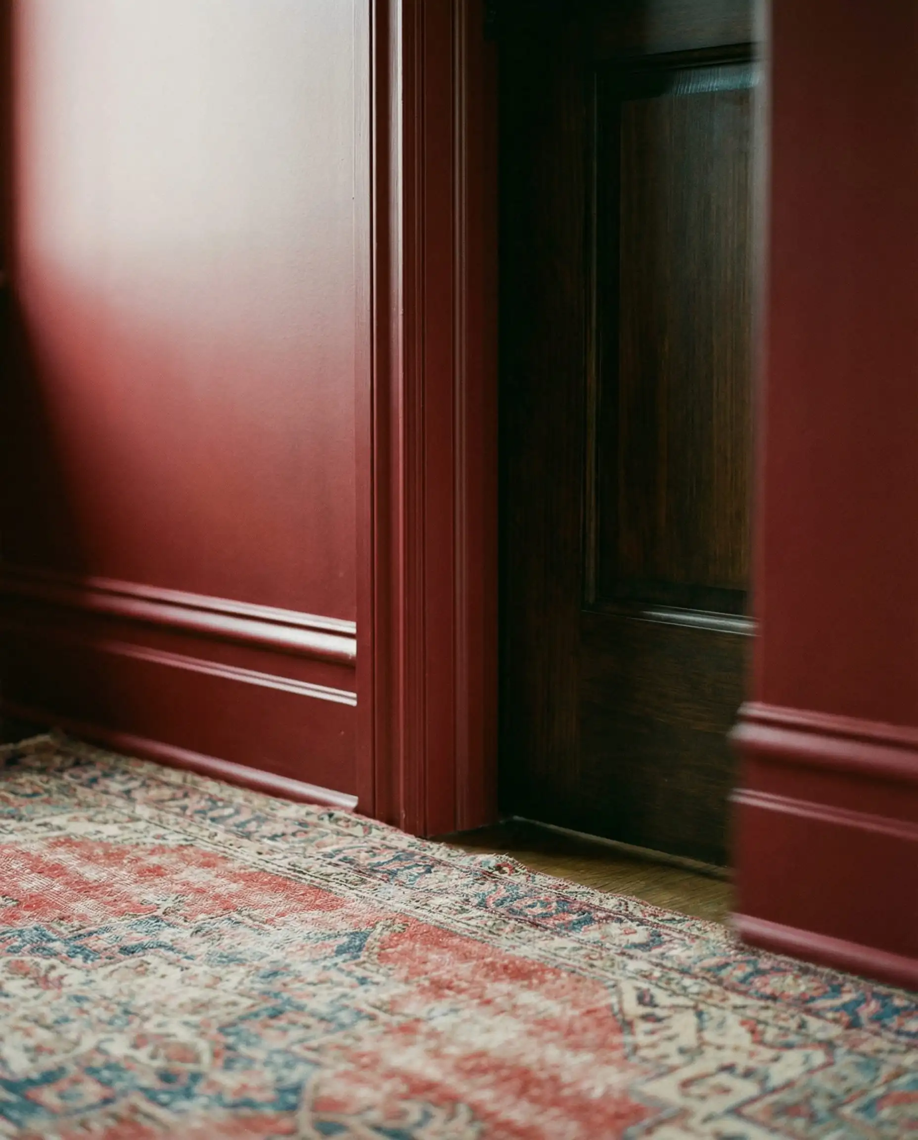 A detail shot focusing on a door frame and baseboard 'Color Drenched' in the same deep, moody red as the wall. This interior features Deep muted red (not fire engine), dark wood, and faded color from the rug tones and a cozy, seamless, warm atmosphere.