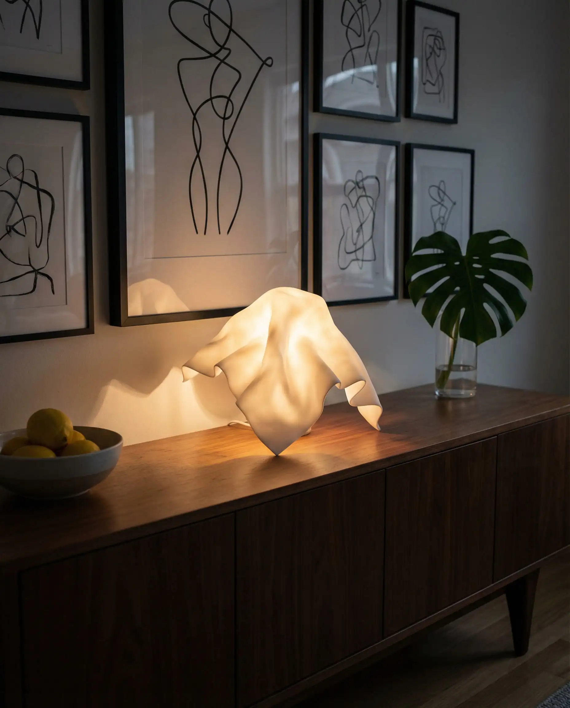 White resin table lamp shaped like a draped napkin or handkerchief, glowing softly on a walnut sideboard.