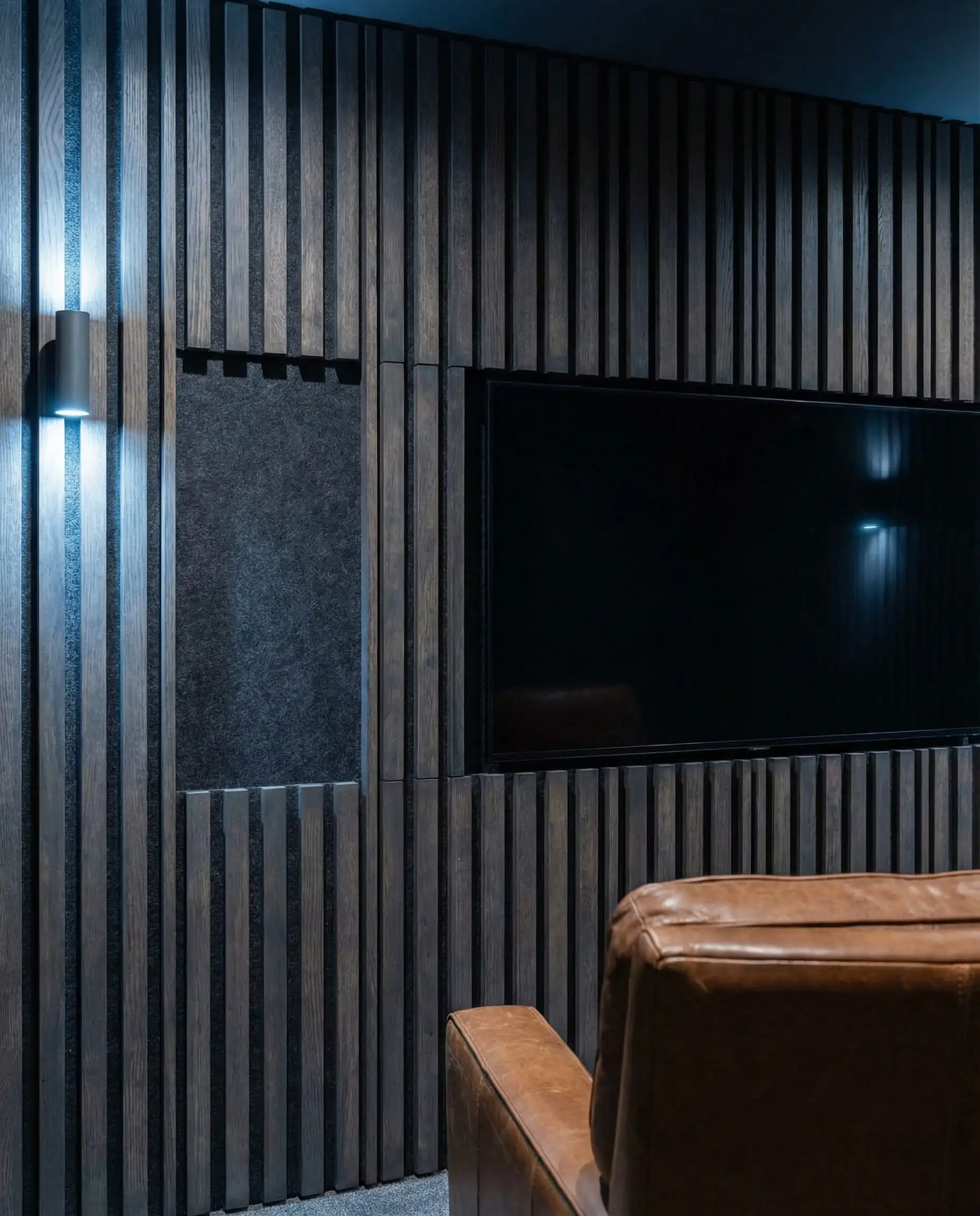 A modern media area where a wall of mixed-width charcoal-stained wood slats is used to camouflage a wall-mounted TV. This media area features Charcoal grey, matte black felt, and rich cognac leather tones and a functional luxury, high-tech, moody atmosphere.
