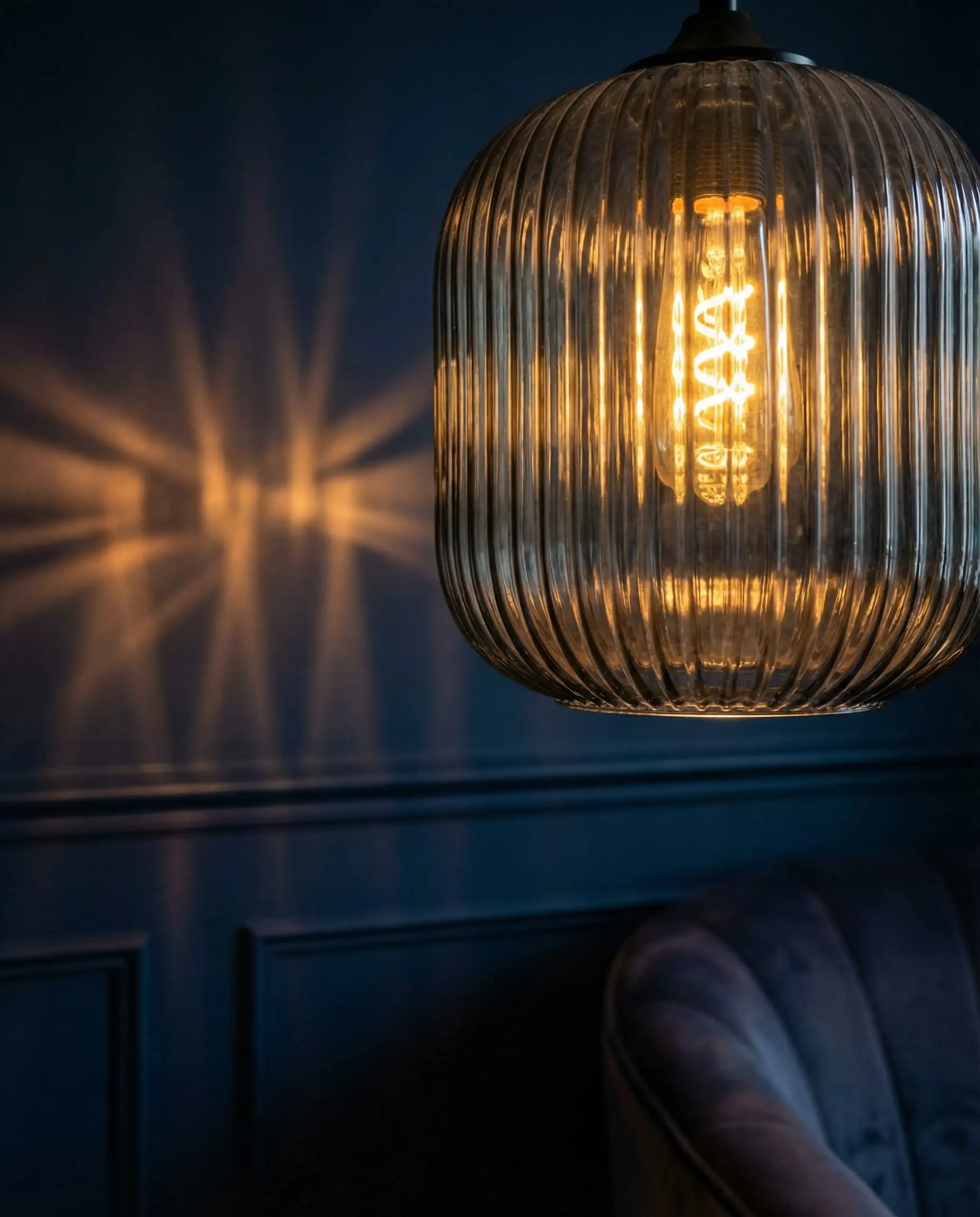 Close-up of a smoked fluted glass lampshade with a vintage filament bulb, casting ribbed shadows on a dark blue wall.