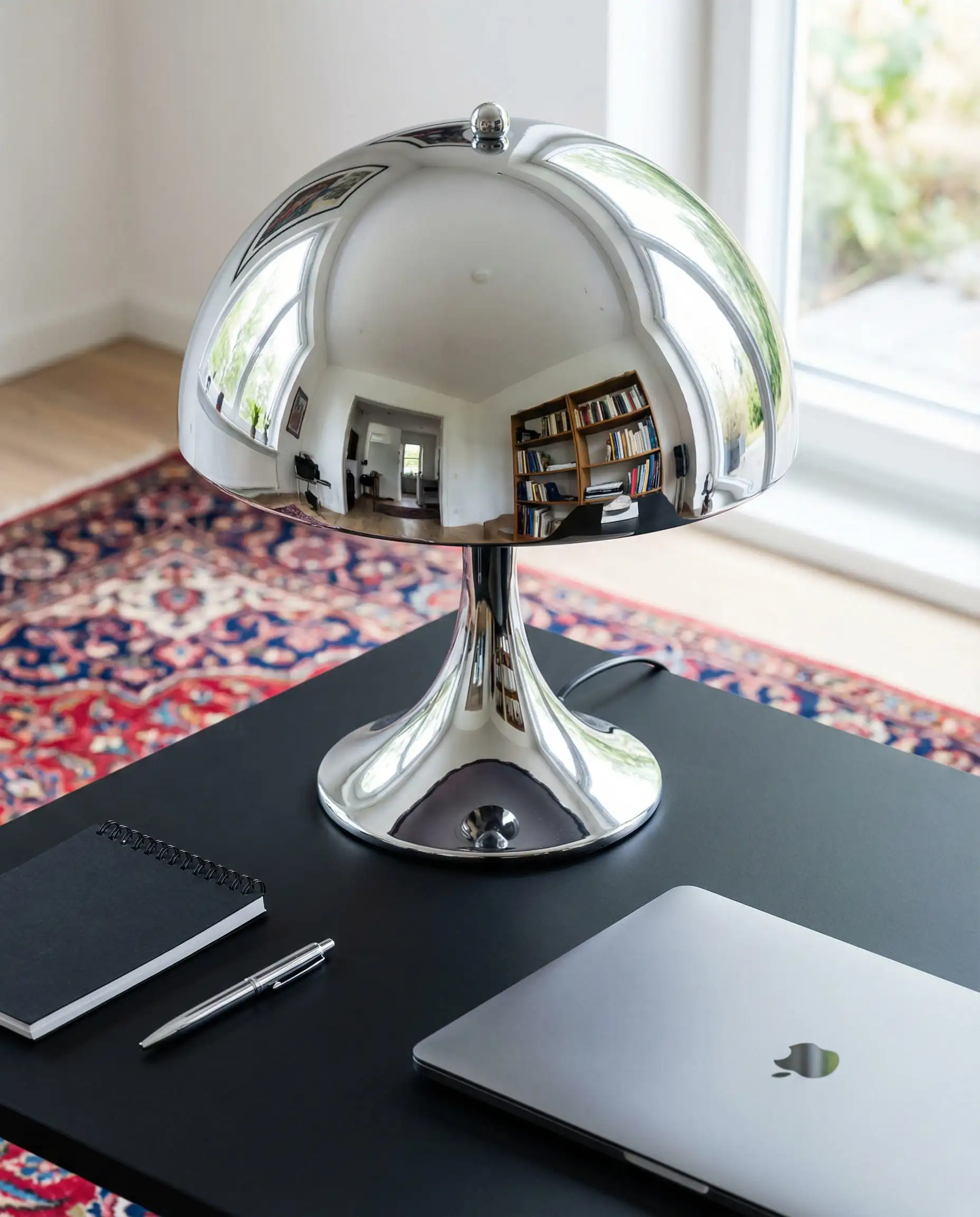 Retro-style high-gloss chrome mushroom table lamp reflecting a colorful living room, styled on a matte black desk.