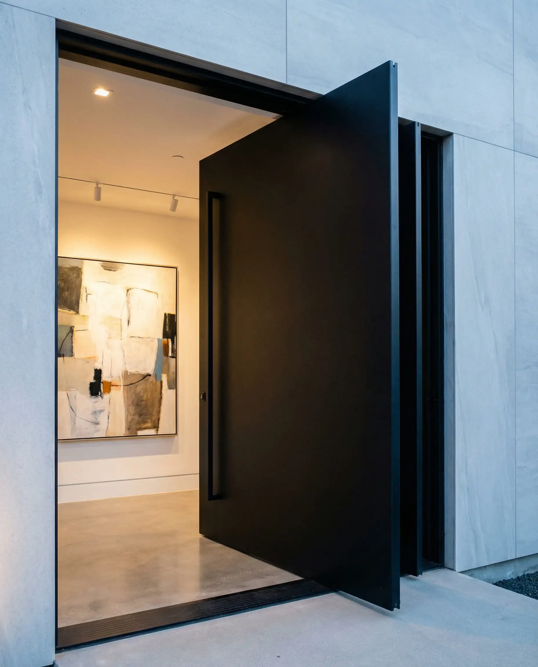 Une grande porte pivotante en aluminium noir mat s'ouvre sur un hall d'entrée moderne en béton.