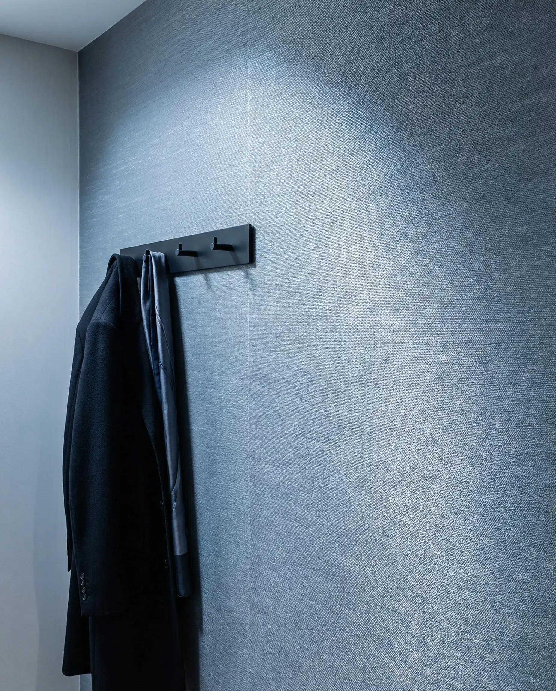 A detail shot of a hallway wall covered in a luxurious, tightly woven silk wallpaper in a soft, silvery-blue tone. This hallway features Silvery blue, matte black, and light grey from the surrounding walls tones and a formal, polished, luxurious atmosphere.