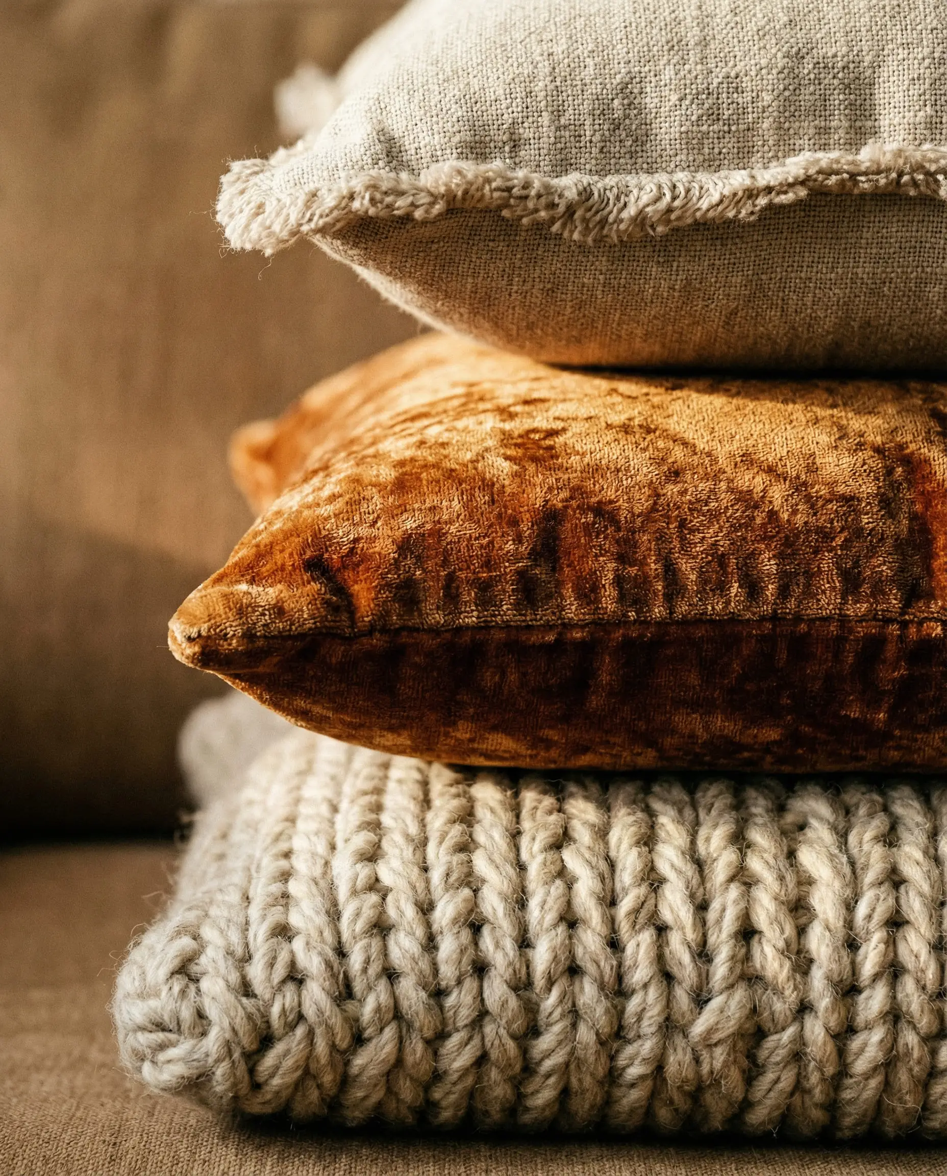 Macro shot of stacked throw pillows showing textures of chunky knit wool, velvet, and raw linen.