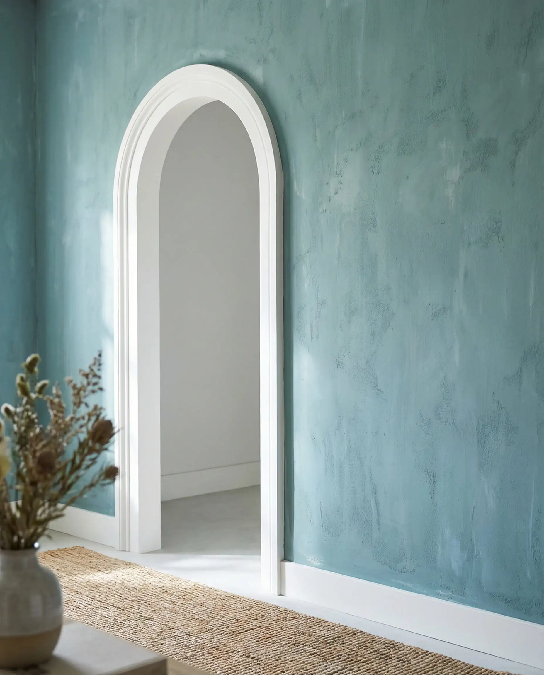 A detail shot of a hallway wall painted in a soft, dusty teal with a chalky, imperfect finish. This hallway features Dusty teal, crisp white trim of the archway, and light grey from the next room tones and a ethereal, soft, tranquil atmosphere.