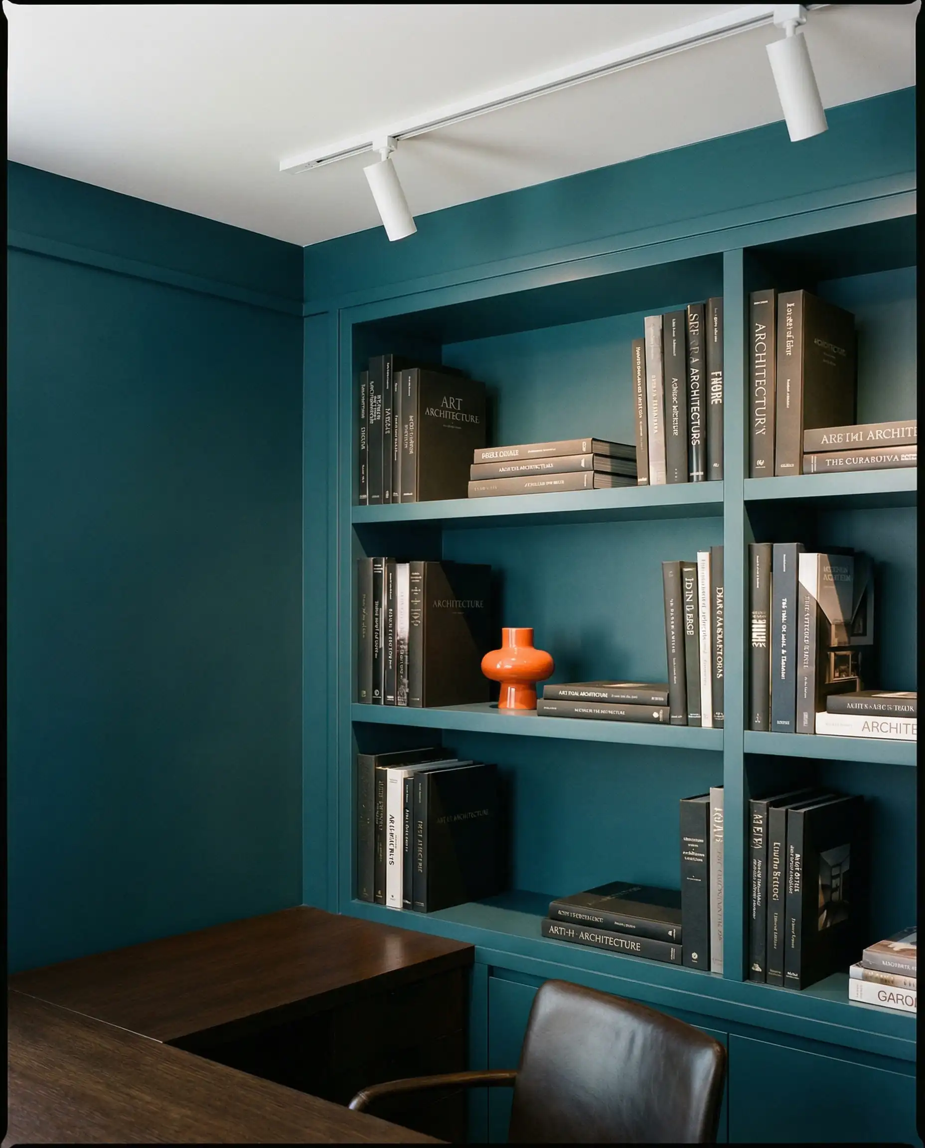 A sophisticated library or home office nook with walls painted in a crisp, dark matte teal. This home office features Dark matte teal, white ceiling, and a single bright orange accent tones and a focused, professional, architectural atmosphere.