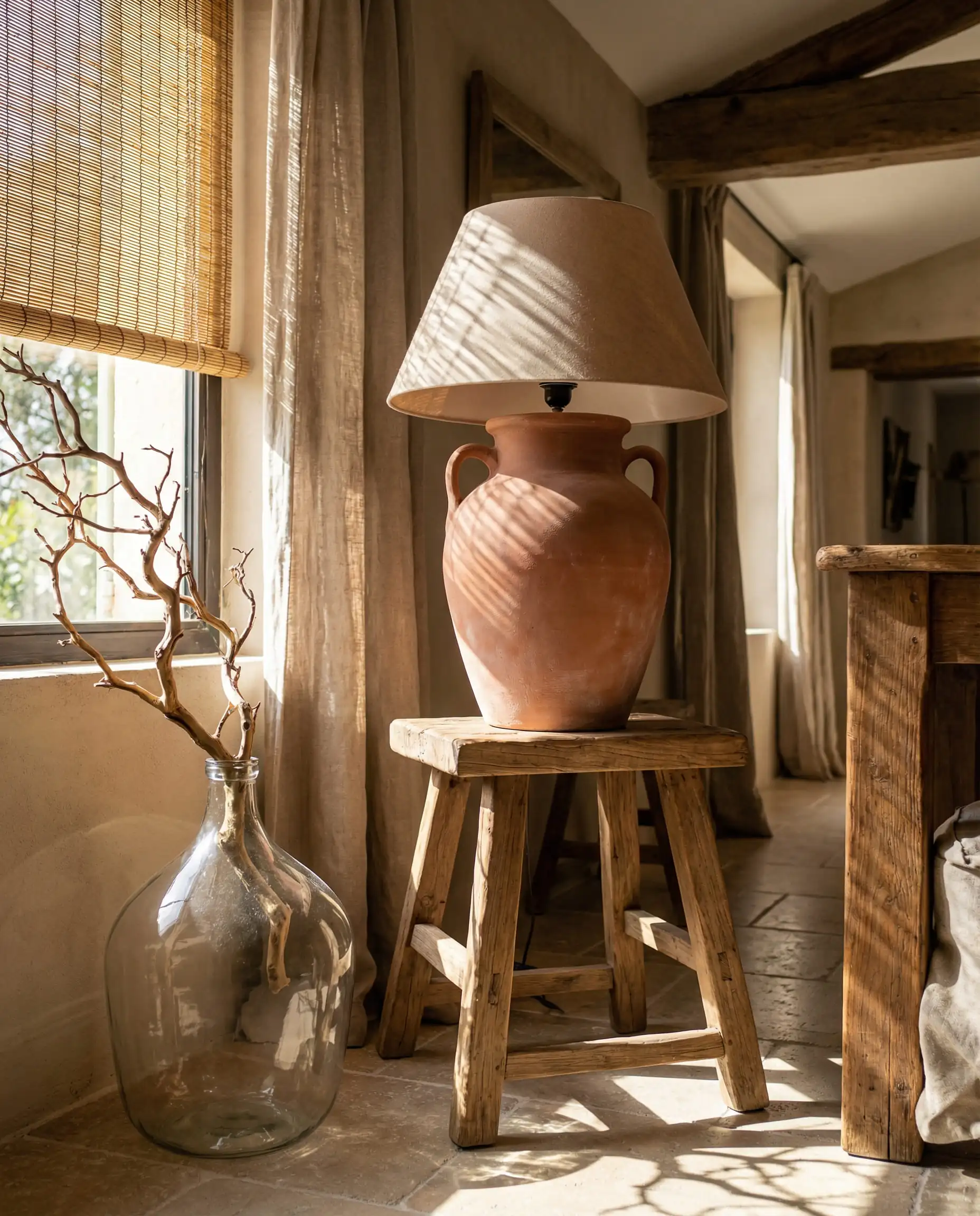 Earthy unglazed terracotta table lamp in a reddish-brown hue, styled on a rustic wooden stool with dried branches.