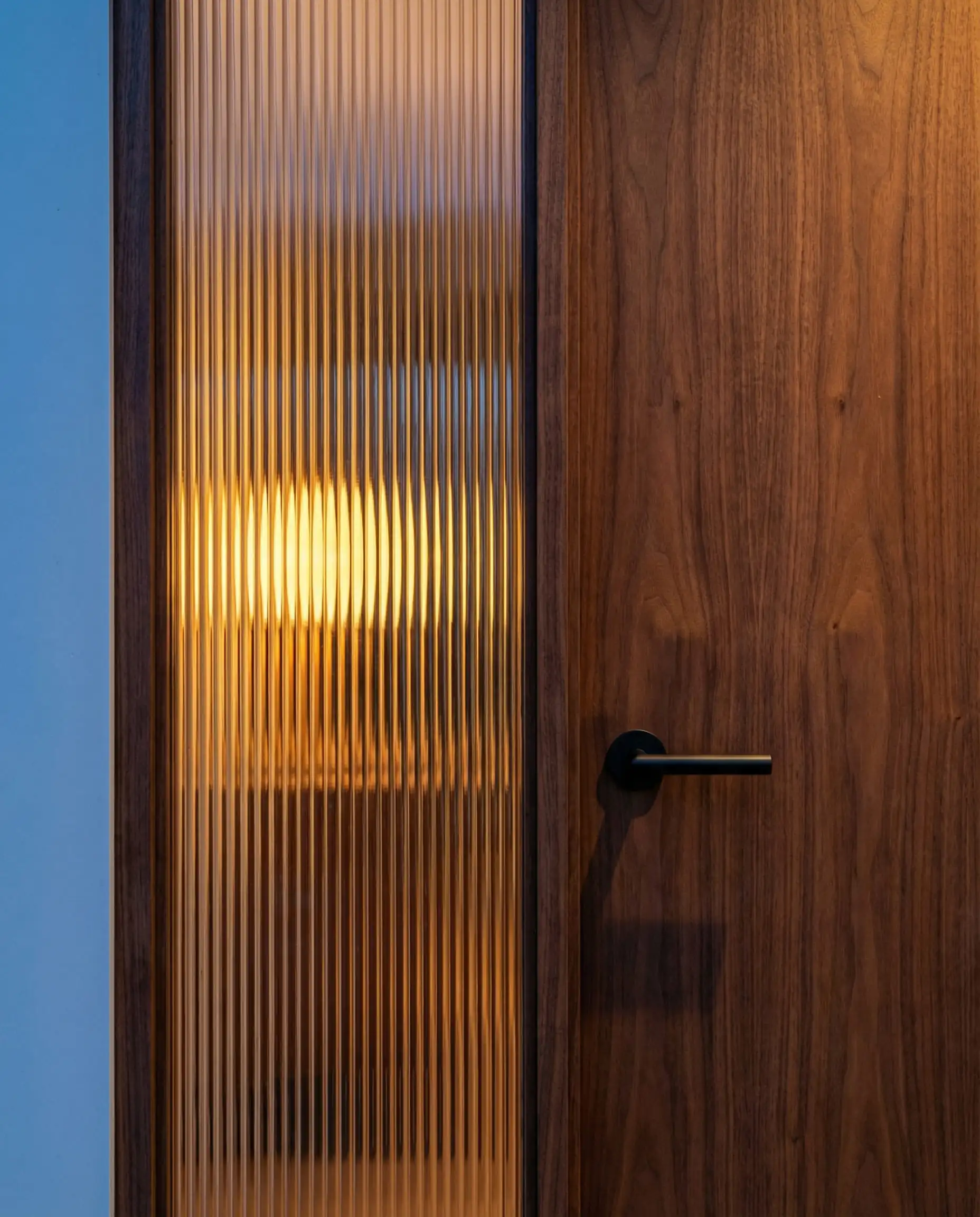 Détail extérieur d'une porte en bois de noyer avec un insert en verre cannelé pour préserver l'intimité, au crépuscule.