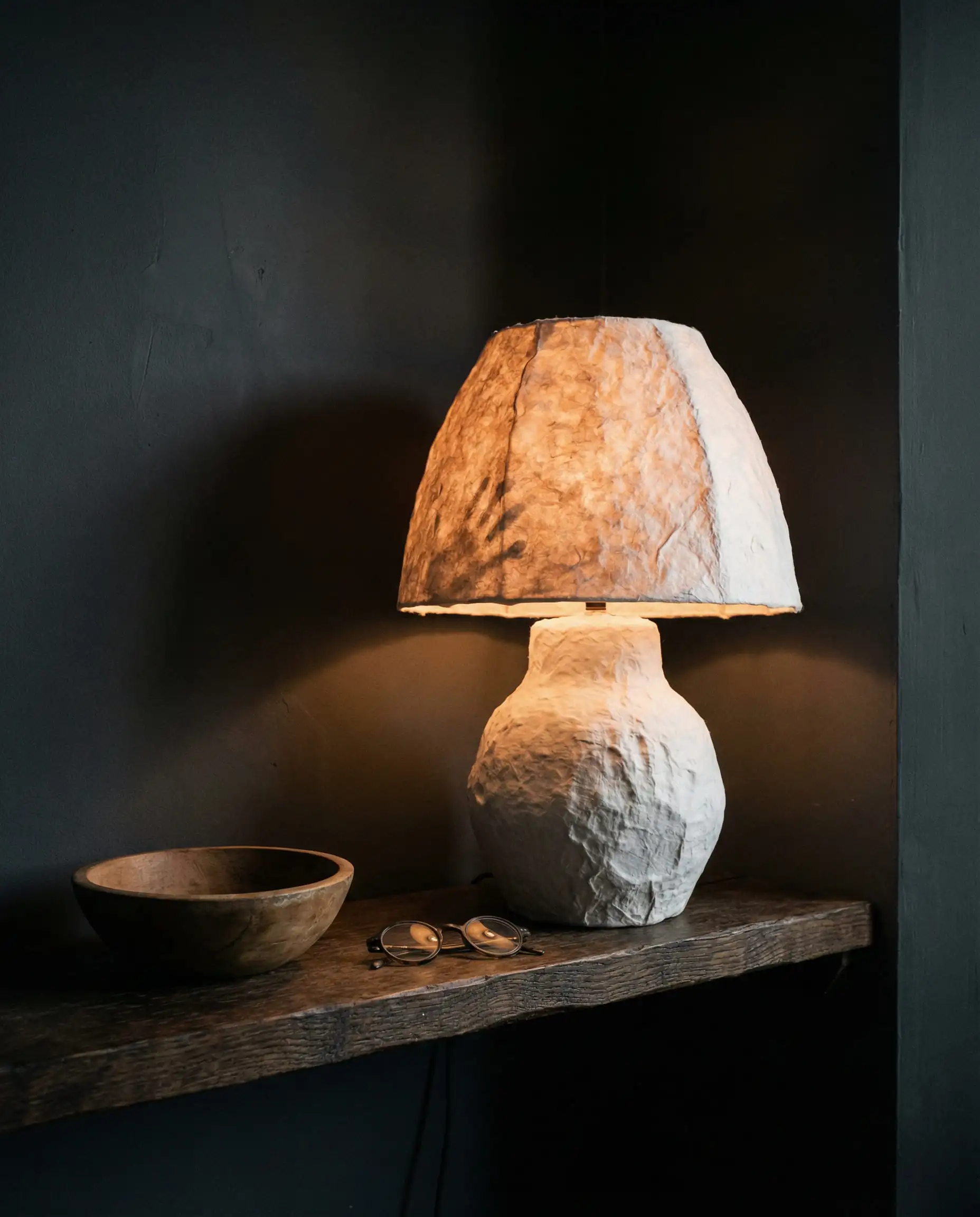 Handcrafted white papier-mâché table lamp with a bumpy organic texture glowing in a dark moody corner.