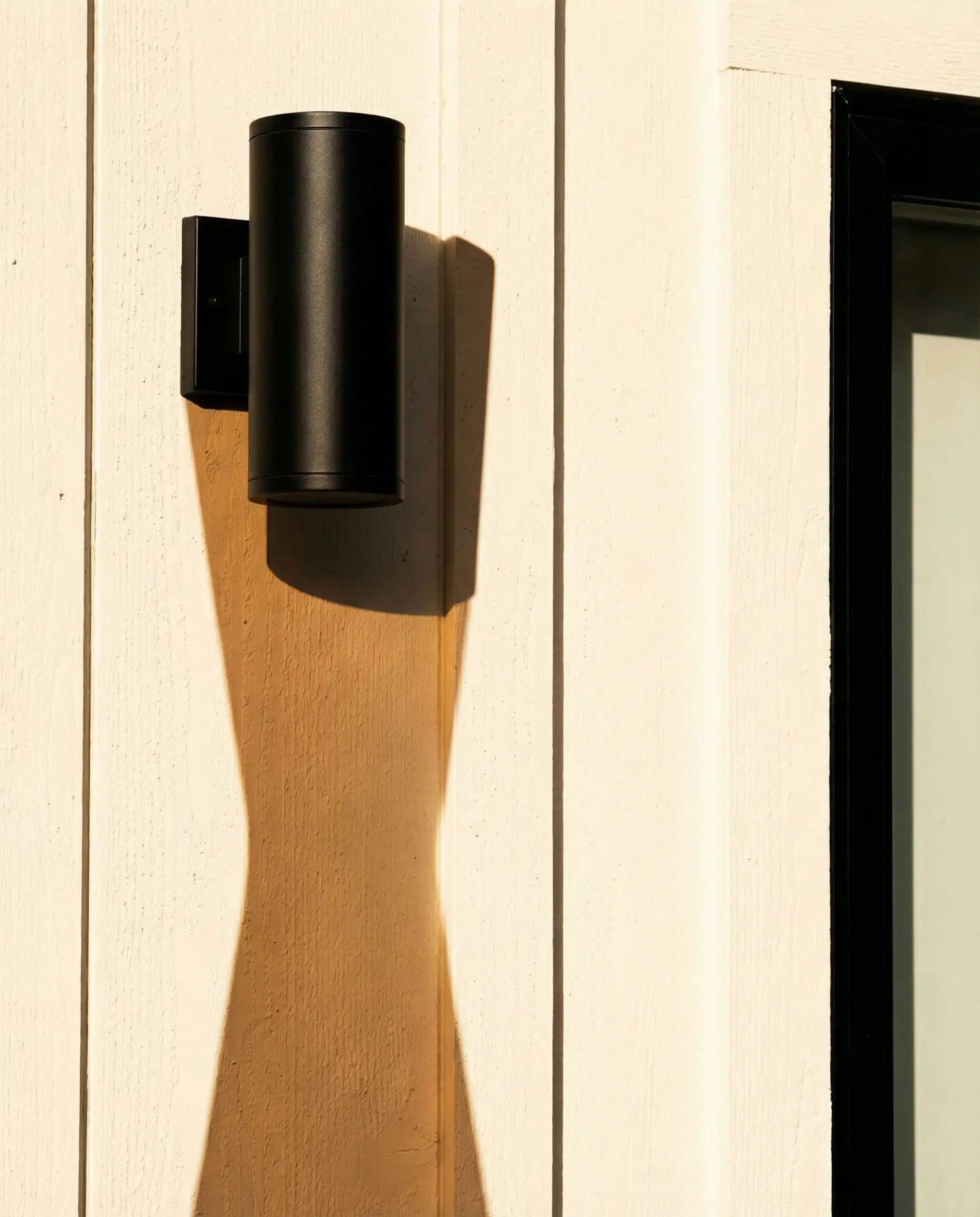 Minimalist detail shot of a matte black modern outdoor wall light casting shadows on vertical siding painted Behr Blank Canvas.