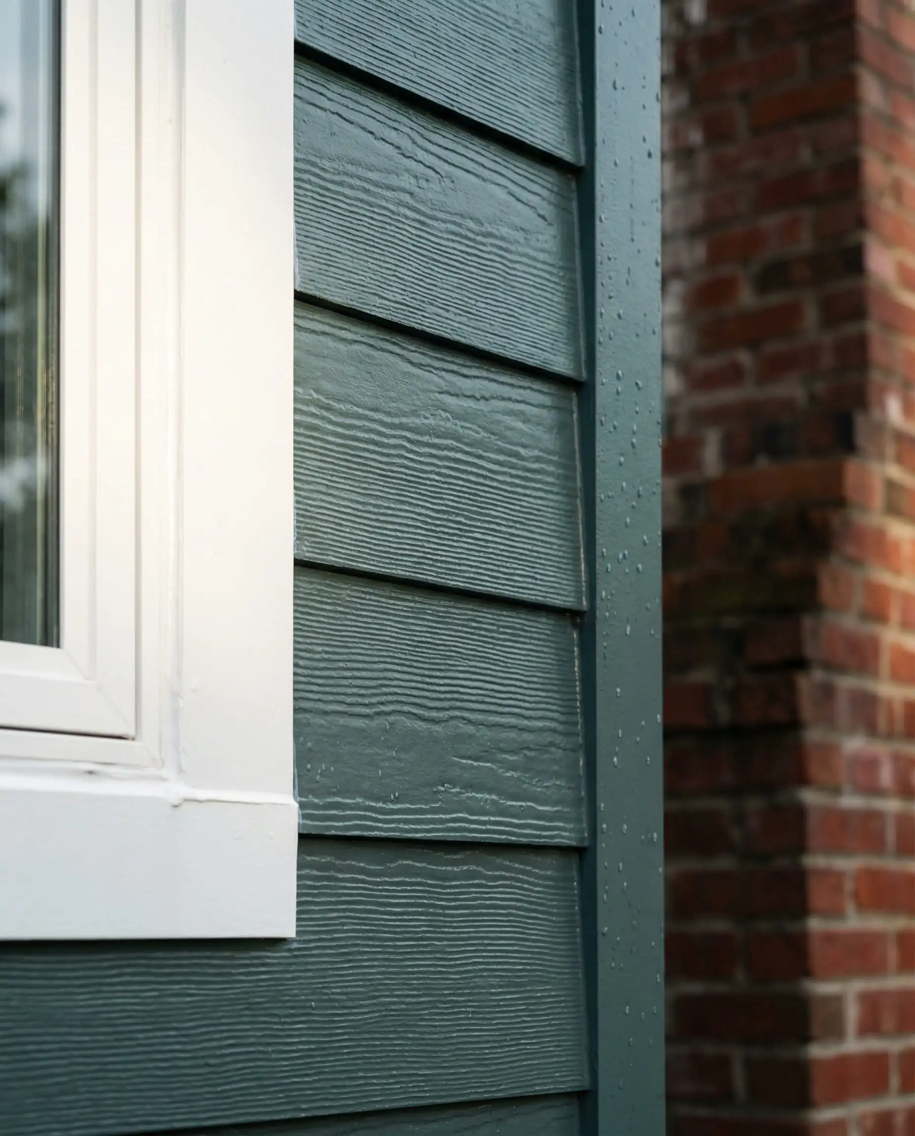Macro detail shot of fiber cement lap siding painted in Behr Hidden Gem showing wood grain texture and white window trim contrast.