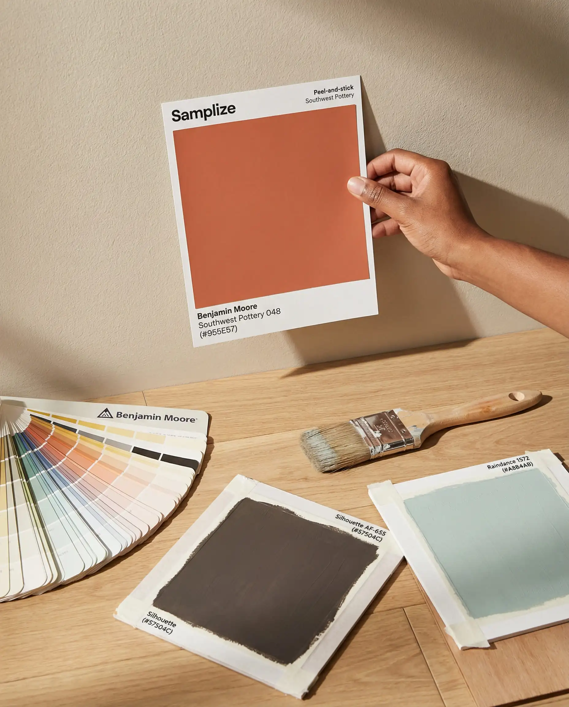 Hand holding peel-and-stick paint samples of Benjamin Moore colors against a wall to test lighting.
