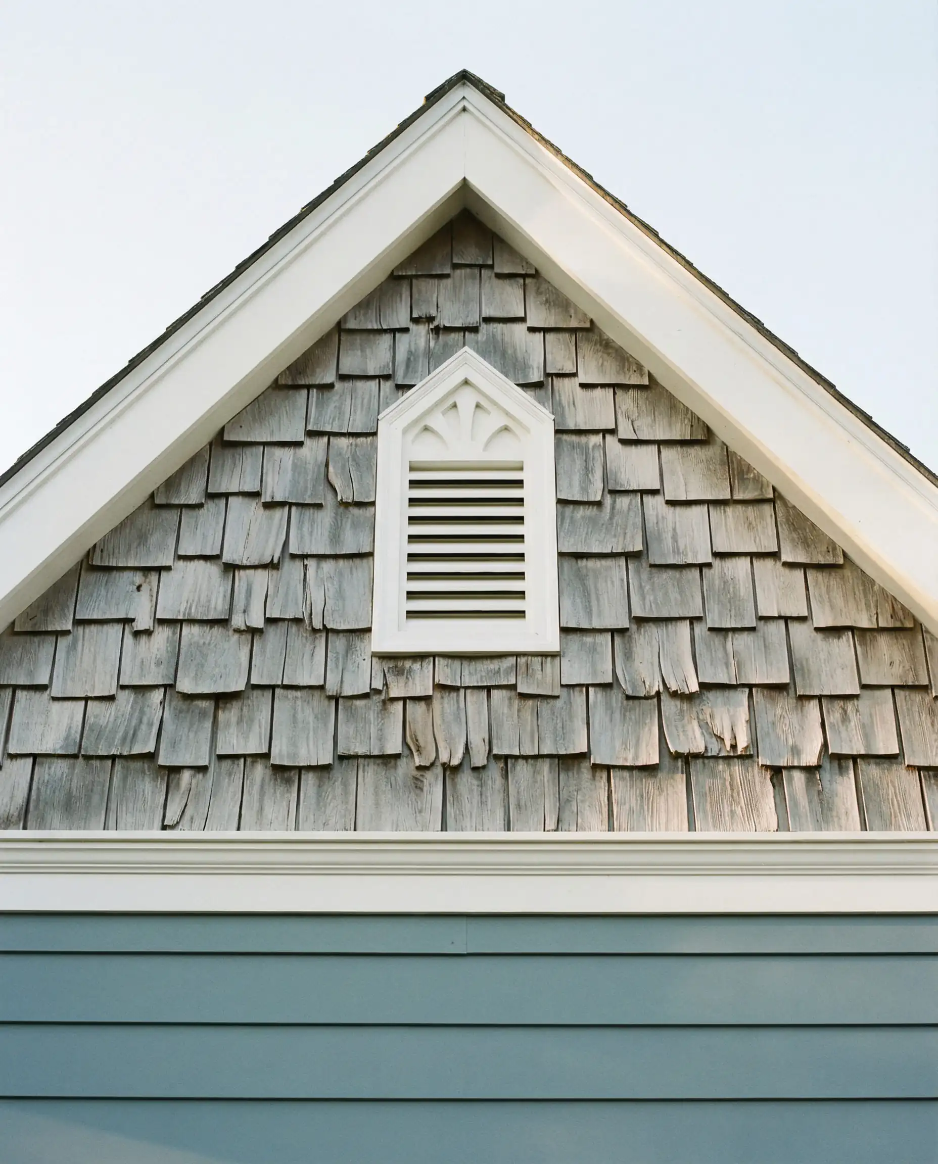 Cottage style gable peak clad in weathered cedar shake shingles sitting above blue horizontal lap siding, separated by white trim.