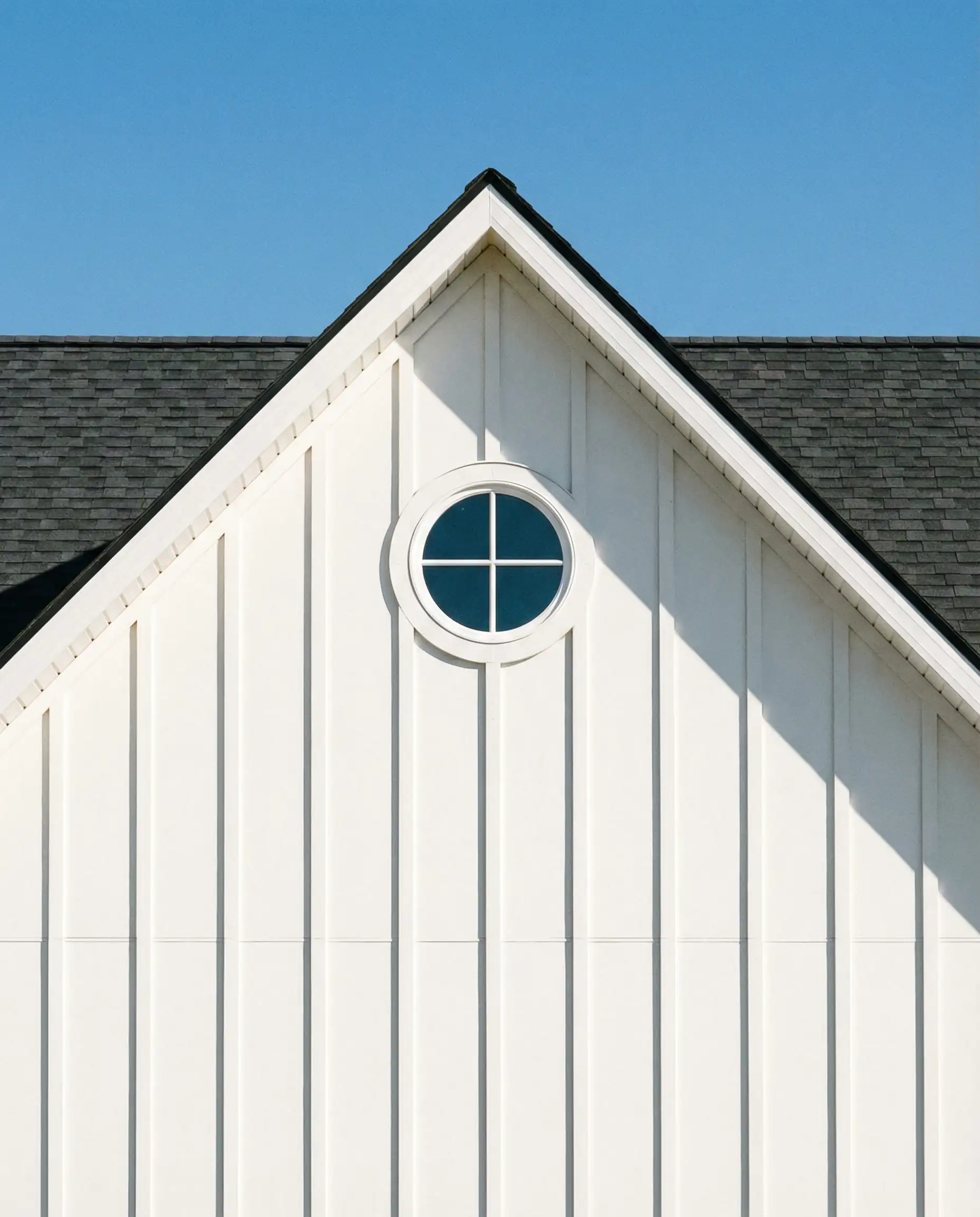 Traditional white farmhouse gable featuring standard width board and batten siding and a round attic window against a blue sky.