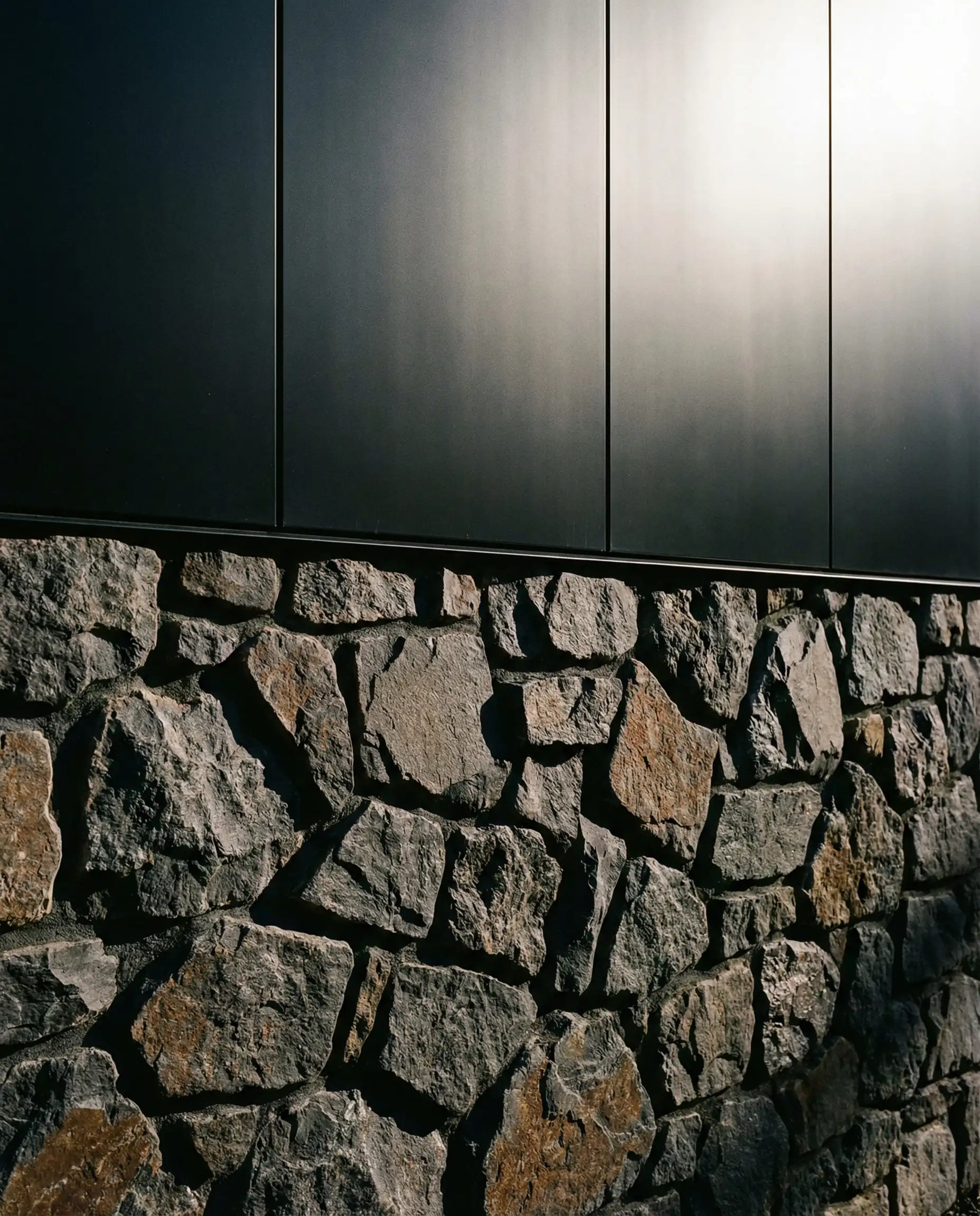 High contrast exterior wall featuring jagged natural fieldstone on the bottom half and smooth black metal panels on the top half.
