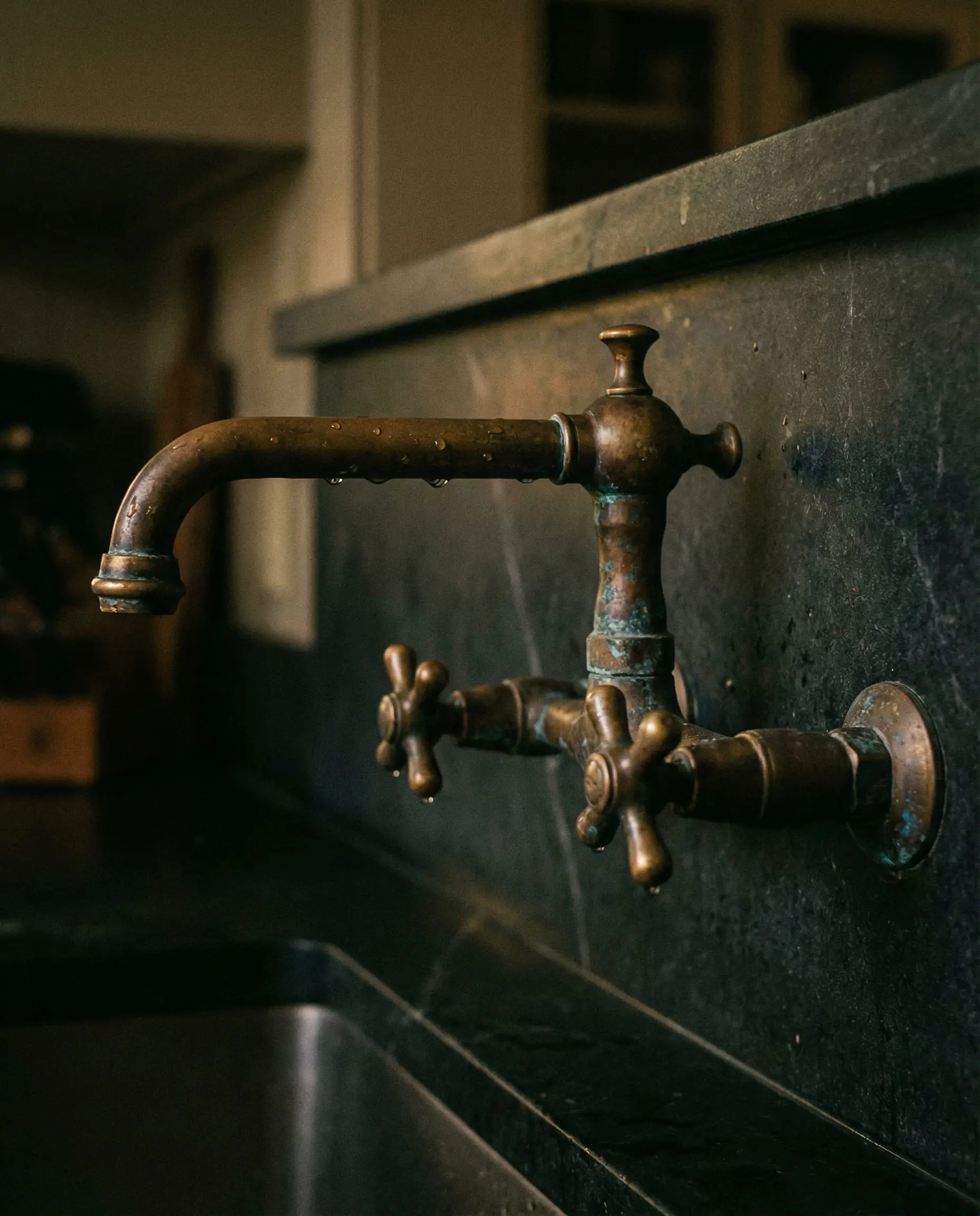 Vintage-style brass faucet showing dark oxidized patina and water droplets against a soapstone backsplash.
