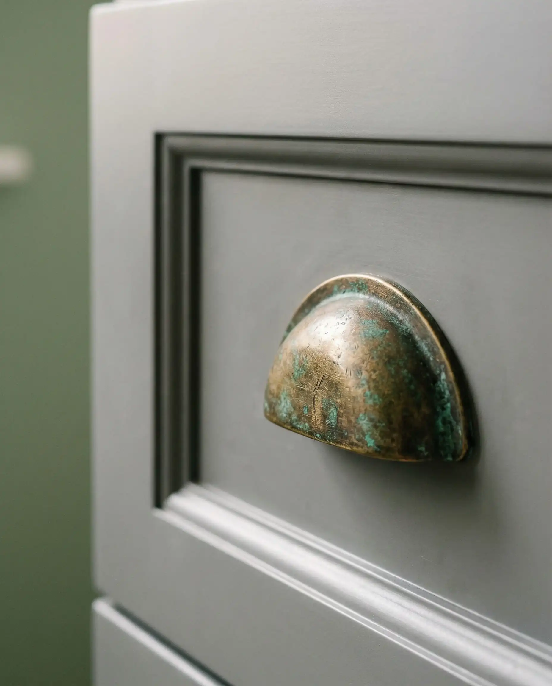 Macro shot of an antique brass cup pull on a gray drawer front with a blurred sage green wall in the background.