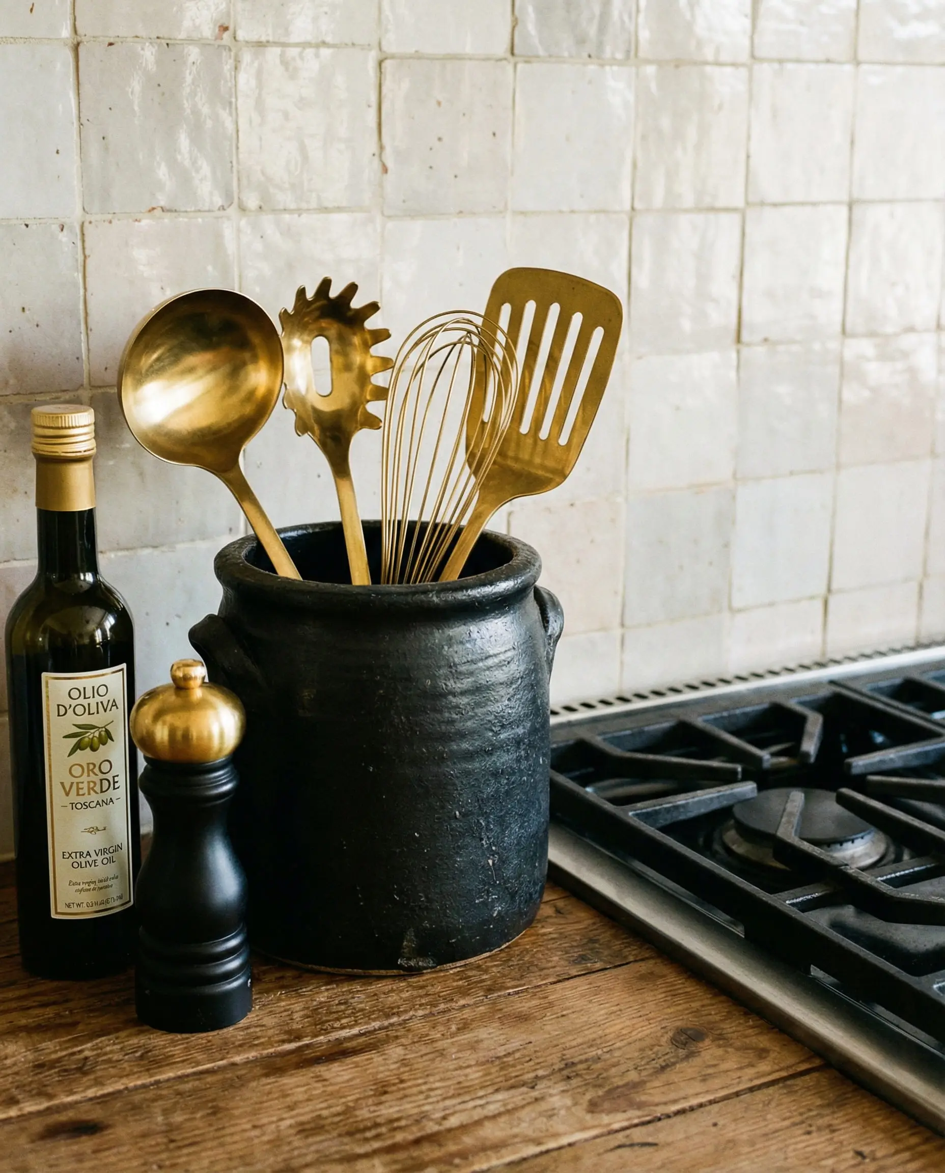 Black ceramic utensil holder filled with gold ladles and whisks next to olive oil.