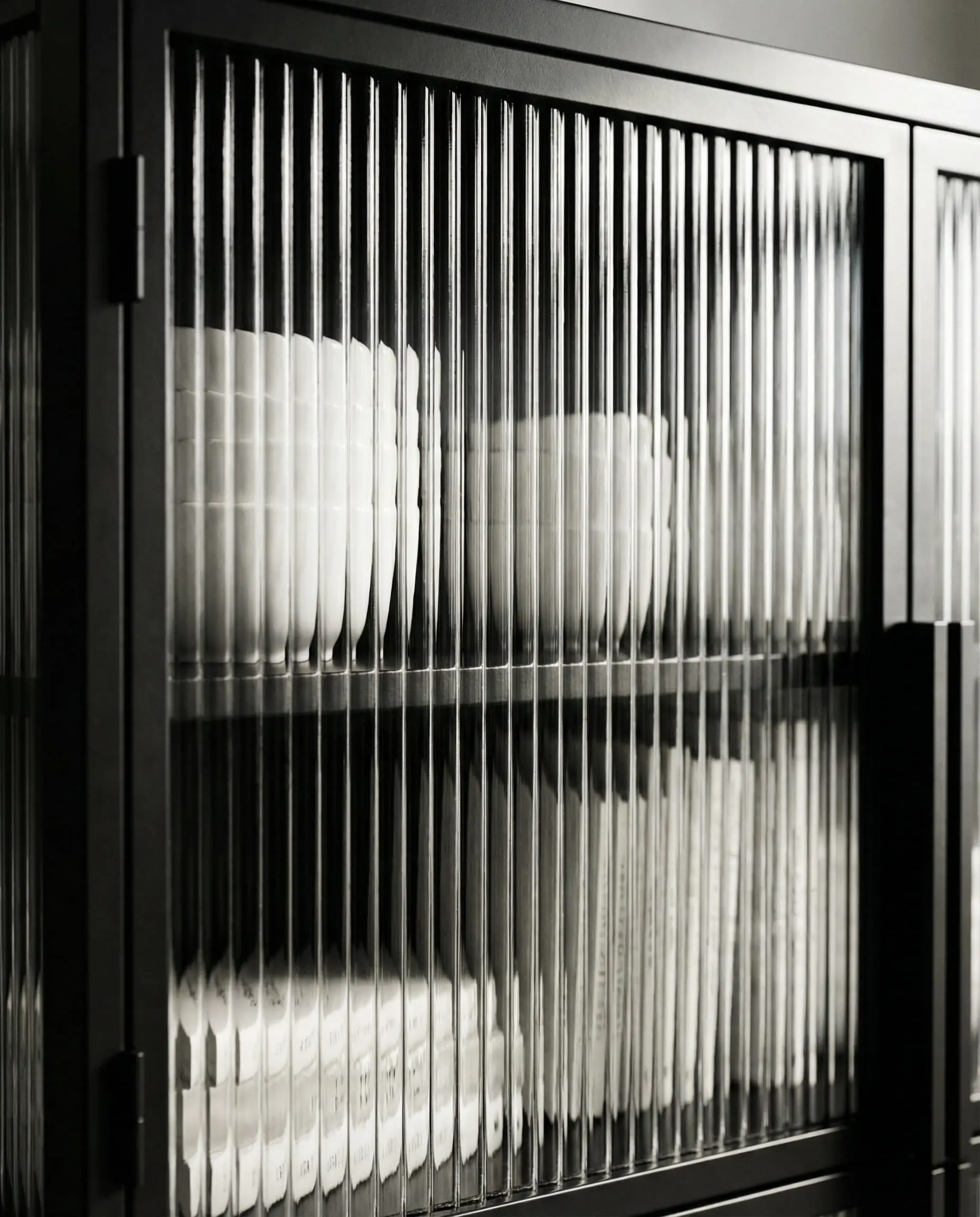 Detail of black metal cabinet with fluted glass doors blurring the dishes and books inside.