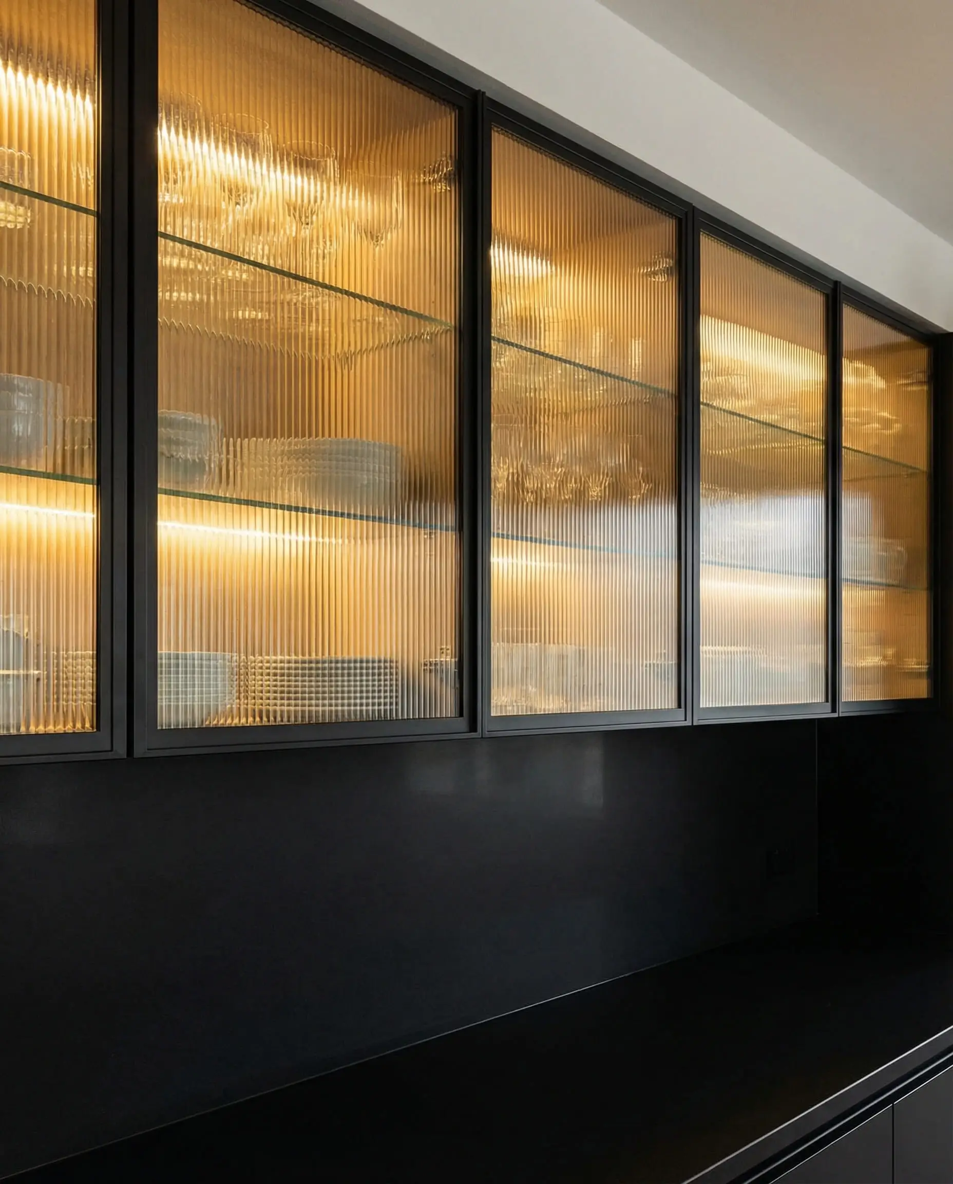 Upper kitchen cabinets with black metal frames and reeded glass inserts illuminated by warm internal LED lighting.