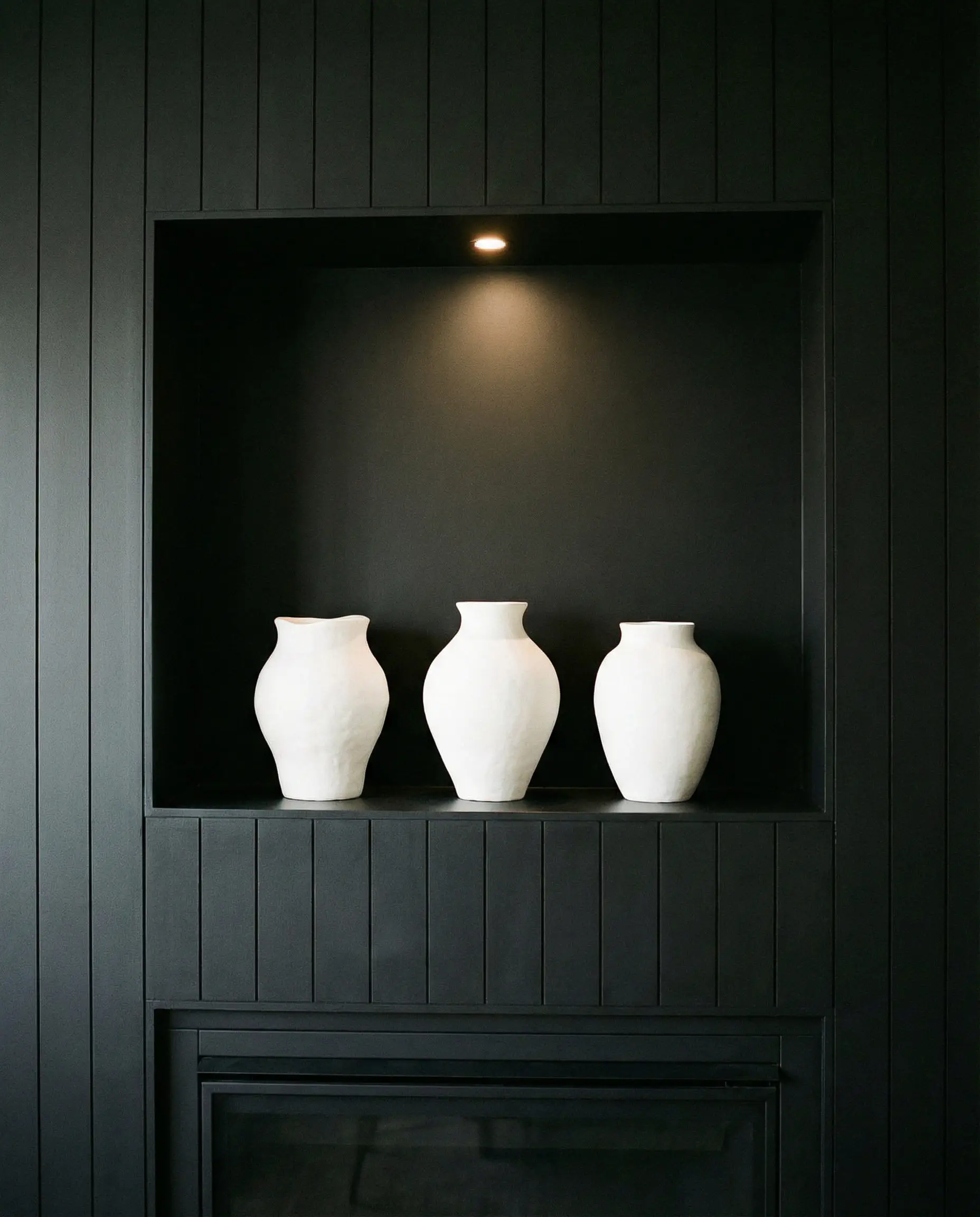 Recessed decorative niche painted black within a black shiplap wall holding white ceramics.