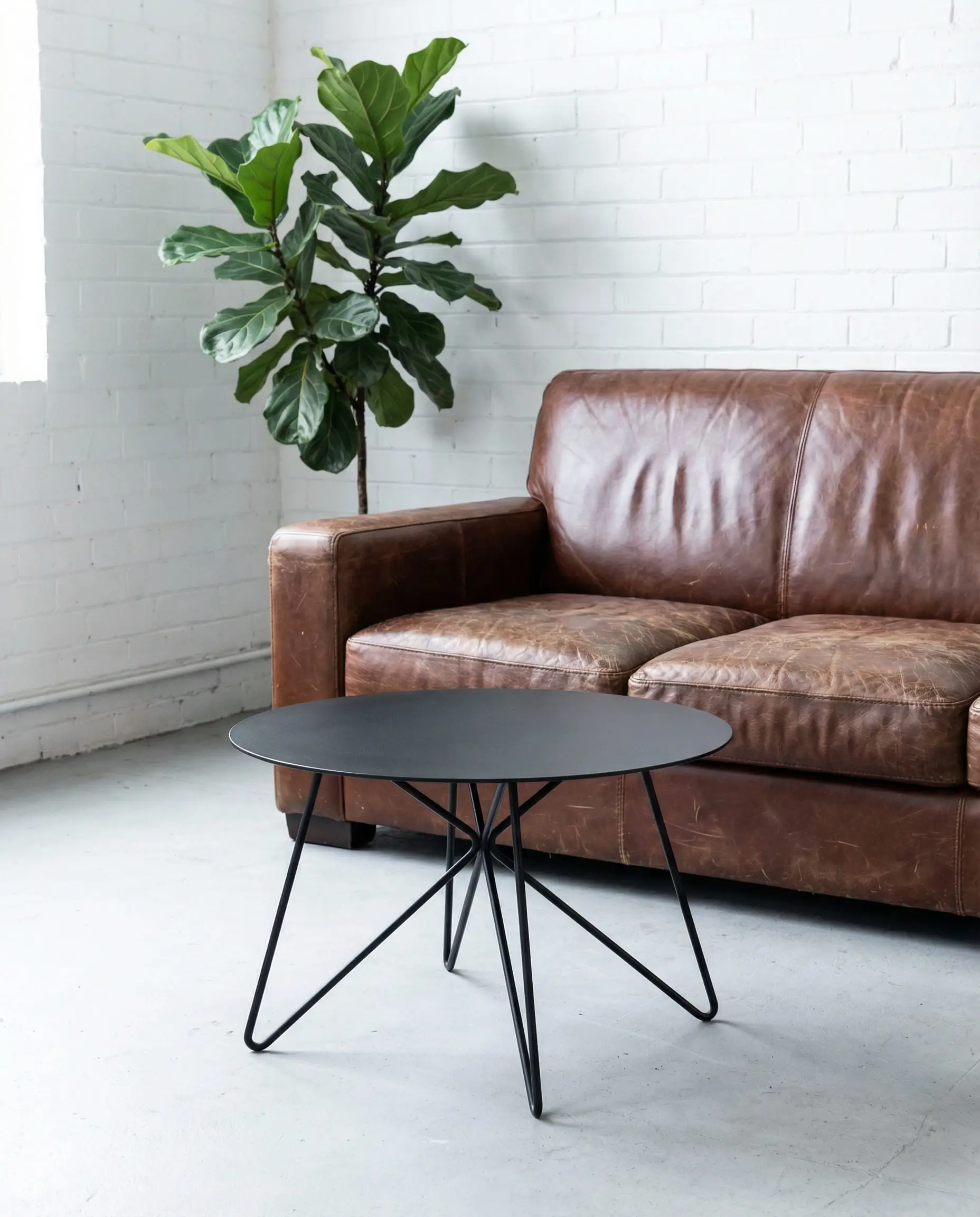 Thin black iron spider-leg coffee table in a minimalist industrial loft setting with a distressed vintage brown leather couch.