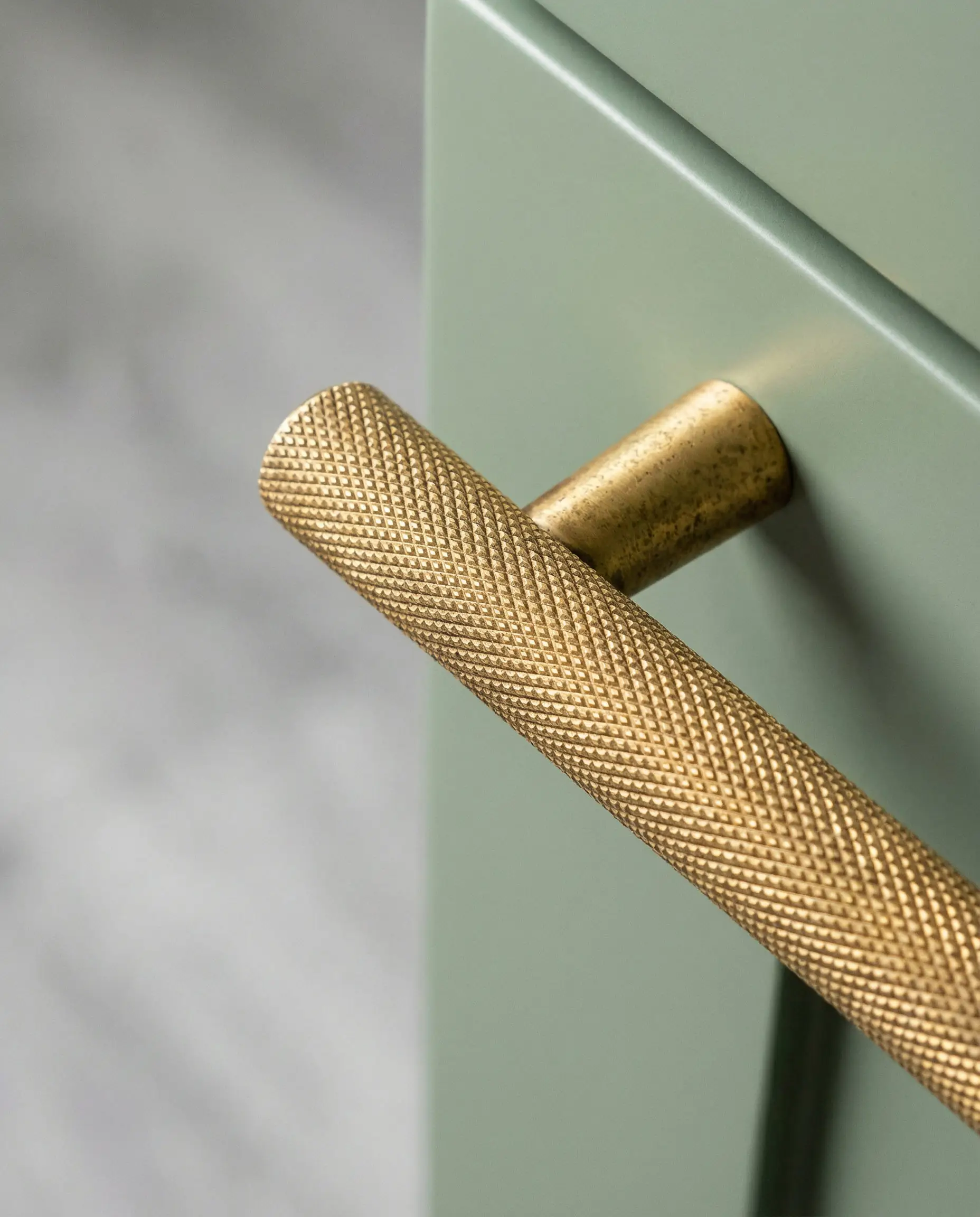 Brushed gold cabinet pull on a sage green door with a blurry gray floor background.