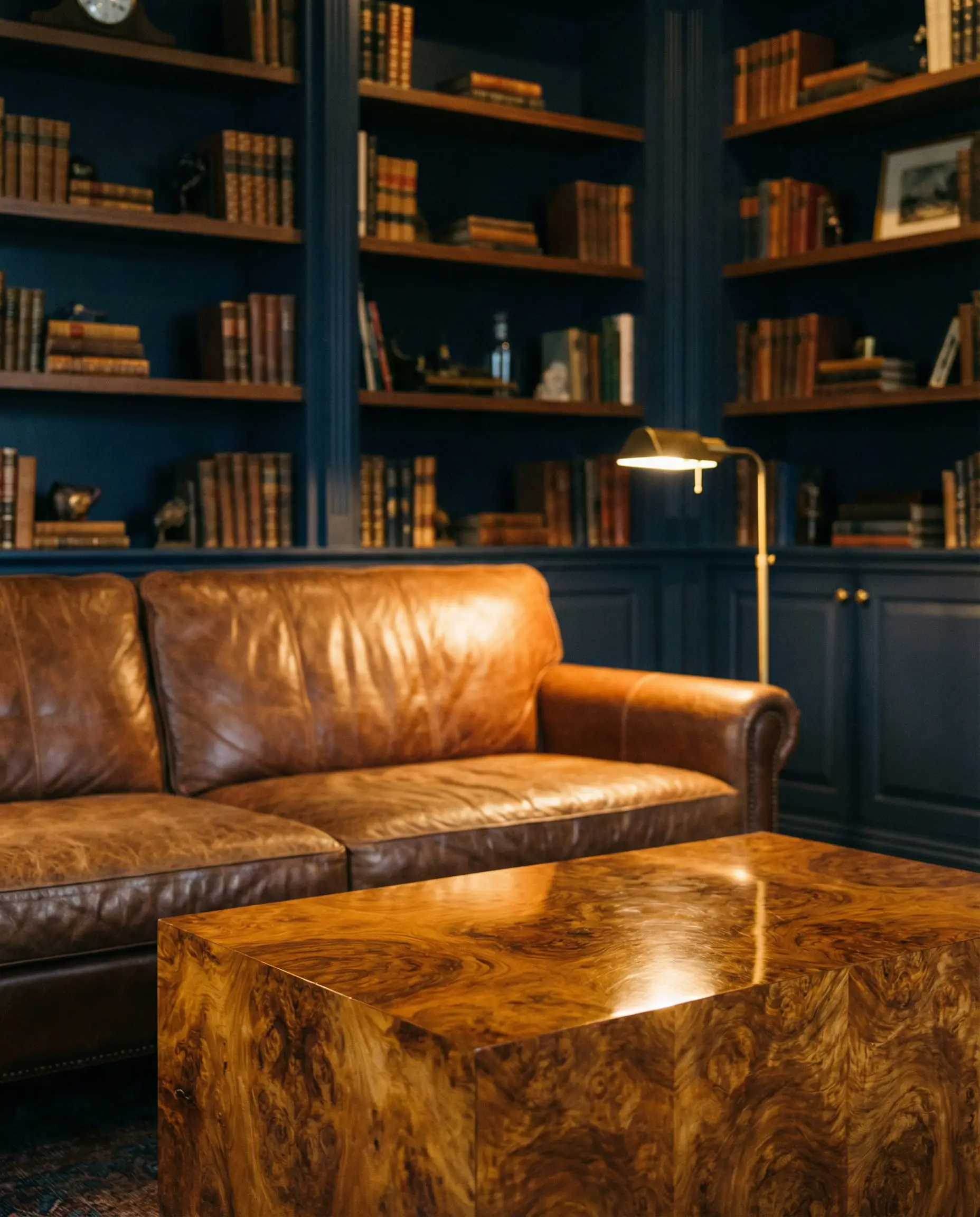 High-gloss block coffee table made of burl wood with swirling grain, positioned in a dark moody library with a saddle brown leather sofa.