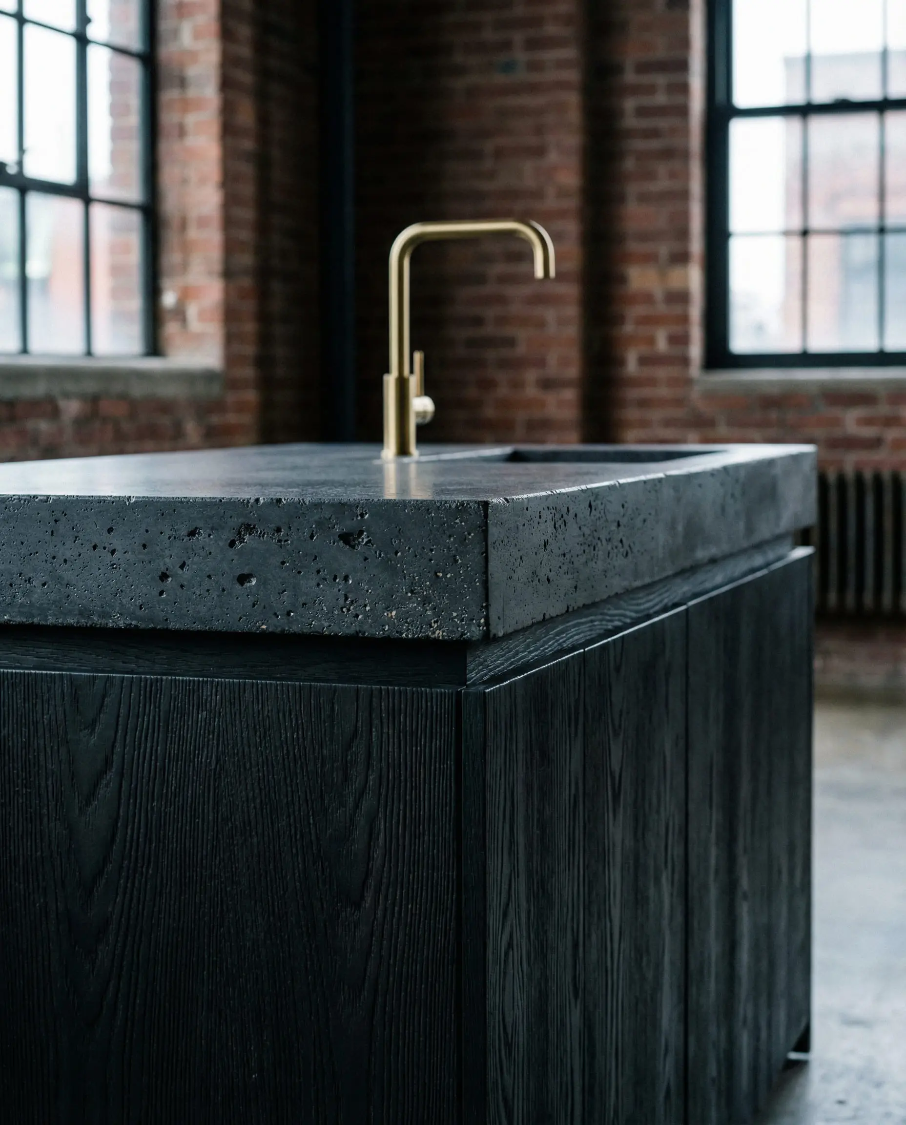 Dark charcoal concrete kitchen island countertop on a black wood base with a minimalist gold faucet.