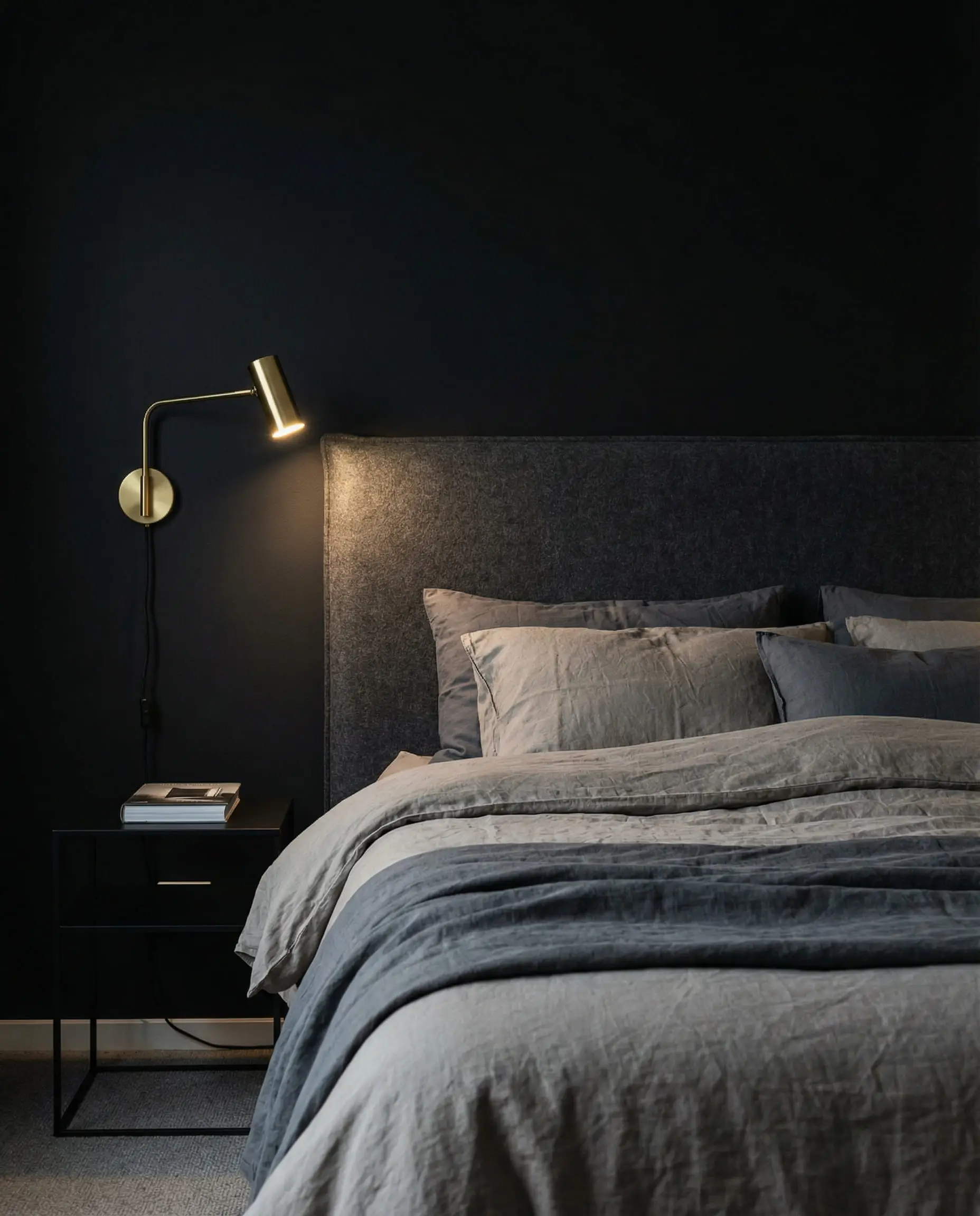 Monochromatic masculine bedroom layering black walls with charcoal wool headboard and greige bedding.