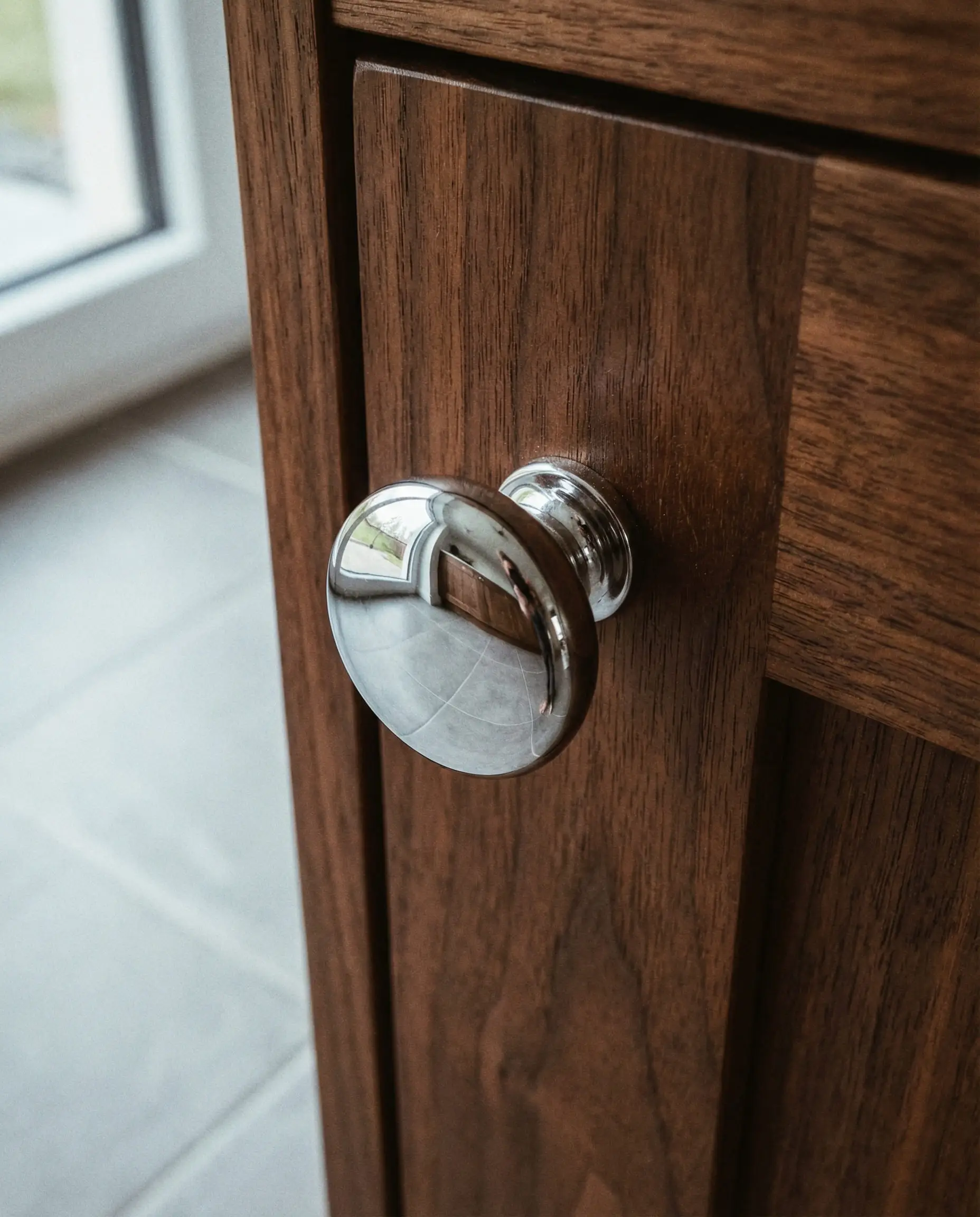 Polished chrome knob on a walnut cabinet door reflecting the gray floor.