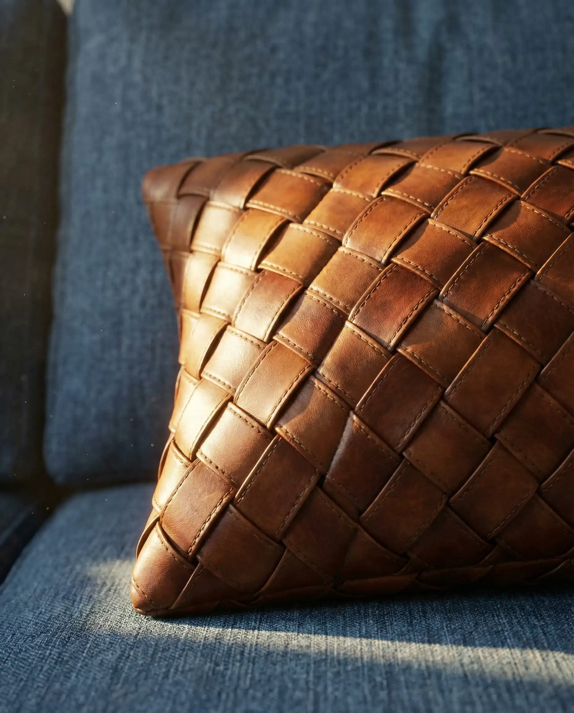 Macro shot of a cognac woven leather pillow resting on a denim blue fabric couch, highlighting the texture contrast.