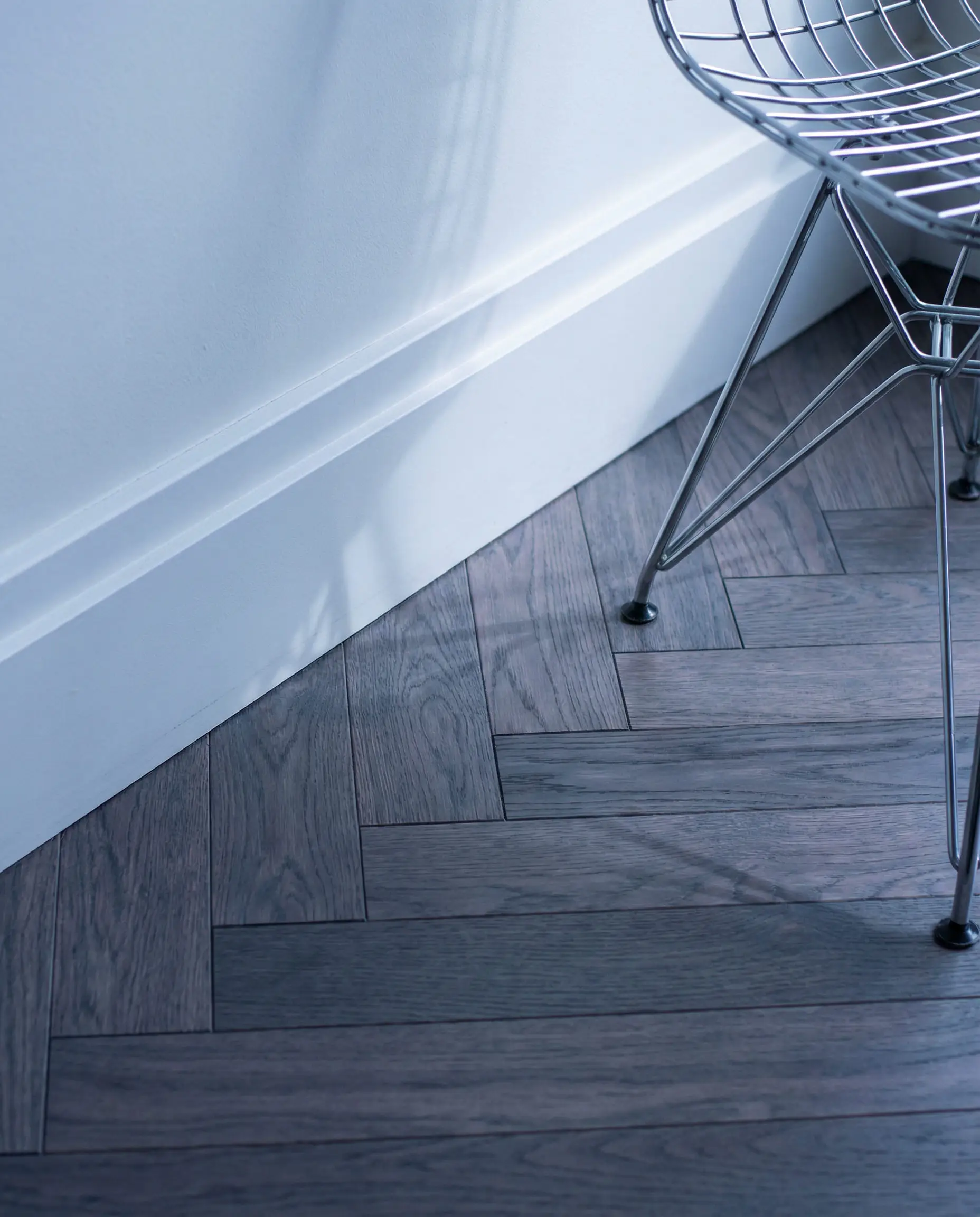 Macro close-up of herringbone gray wood flooring with distinct blue-gray undertones next to a cool white wall and chrome furniture leg.