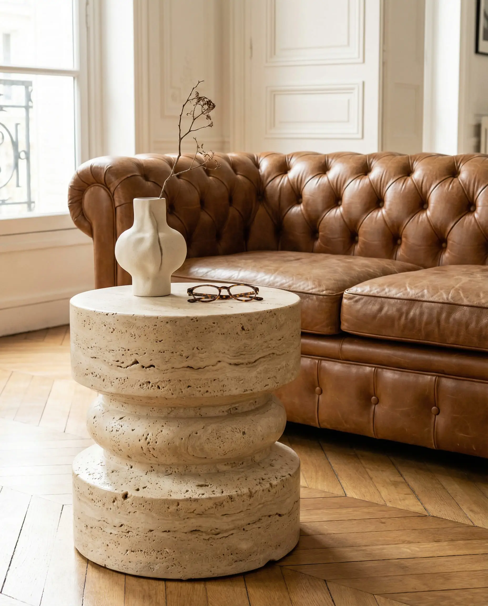 Vertical shot of a chunky cylindrical beige travertine coffee table styled in front of a classic caramel brown leather Chesterfield sofa.