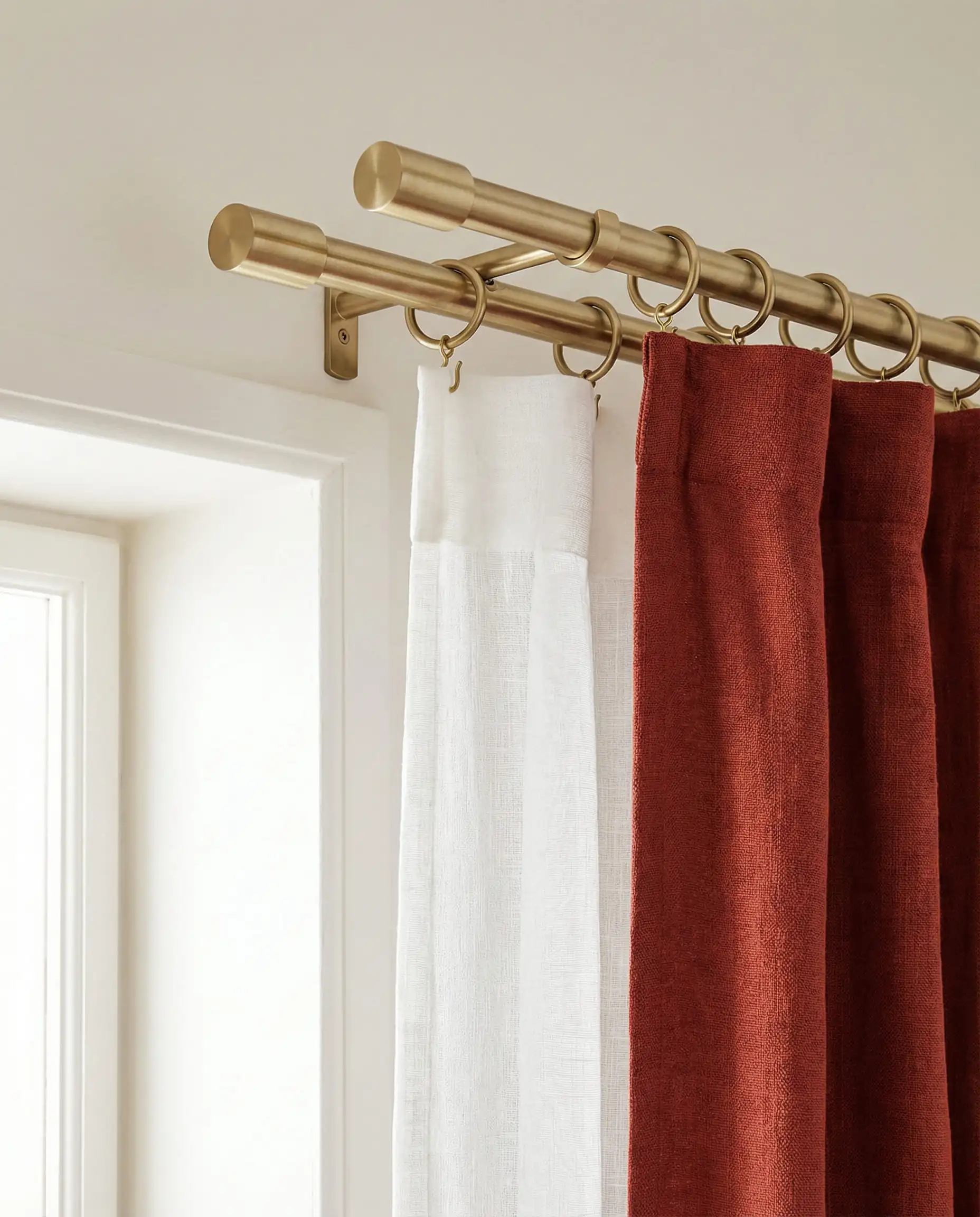 Close-up of a double curtain rod installation showing a white sheer inner curtain and rust red outer drape.