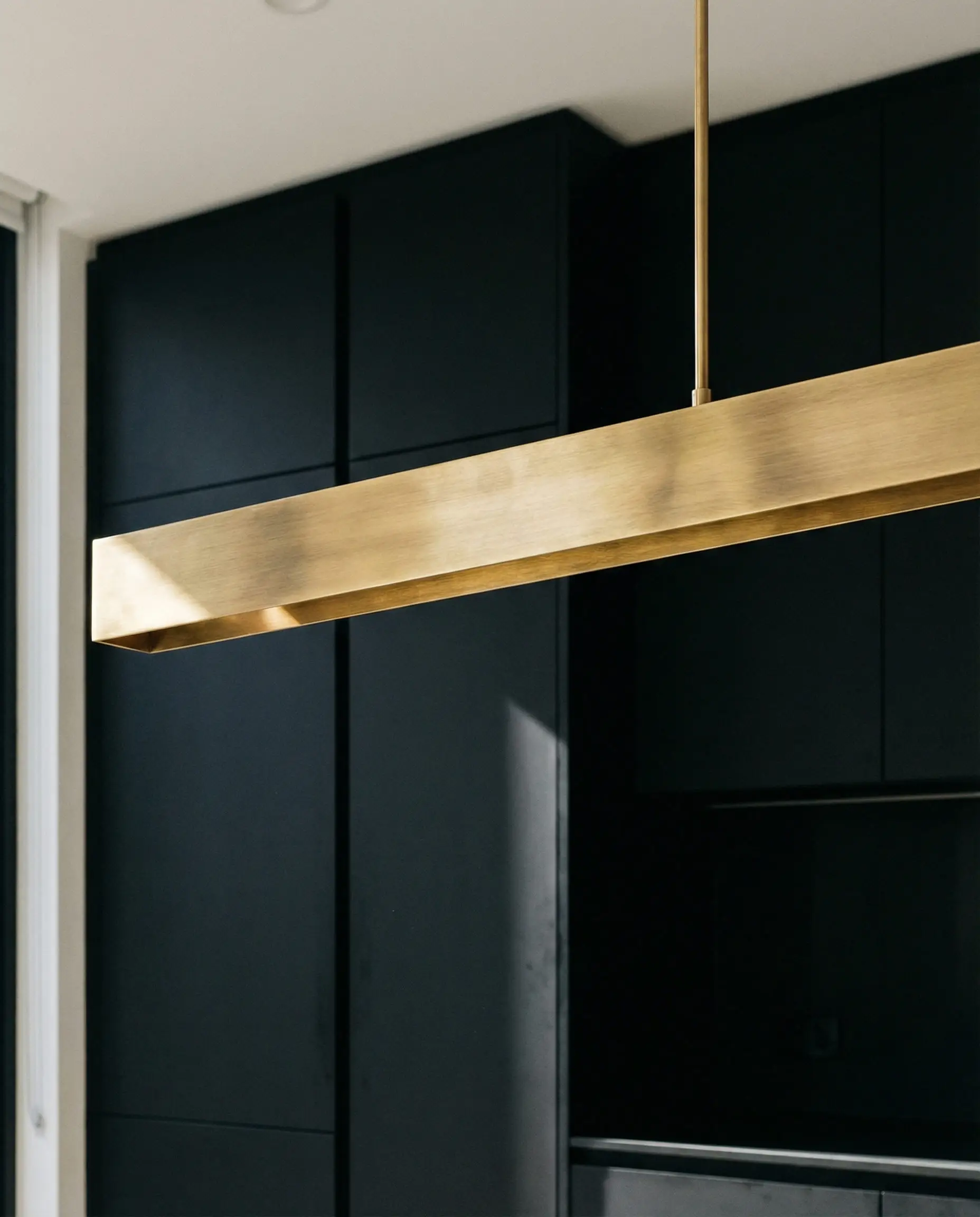 Sculptural brushed brass linear light fixture turned off against a matte black kitchen background.