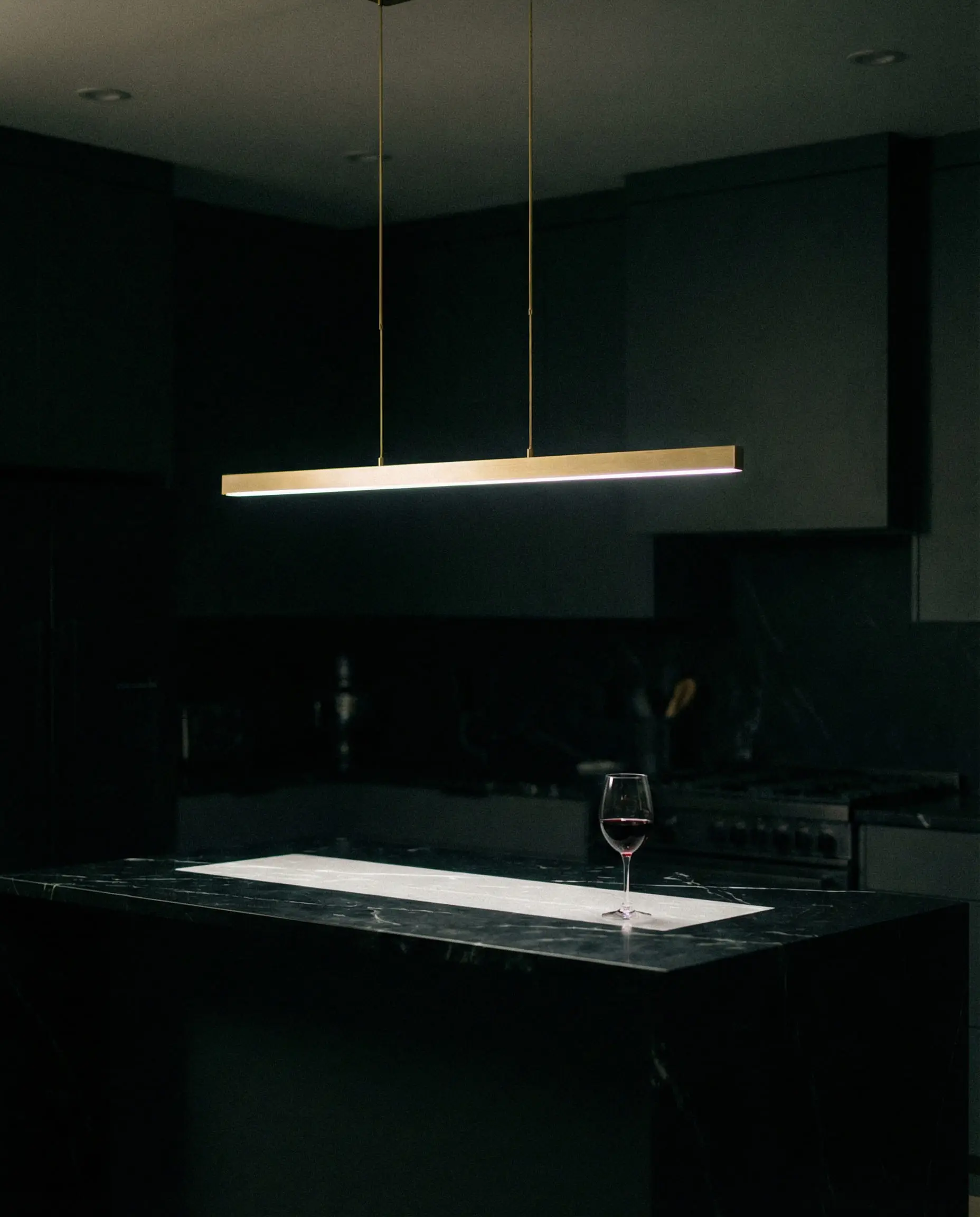 Razor-thin gold LED linear suspension light illuminating a black kitchen island at night.