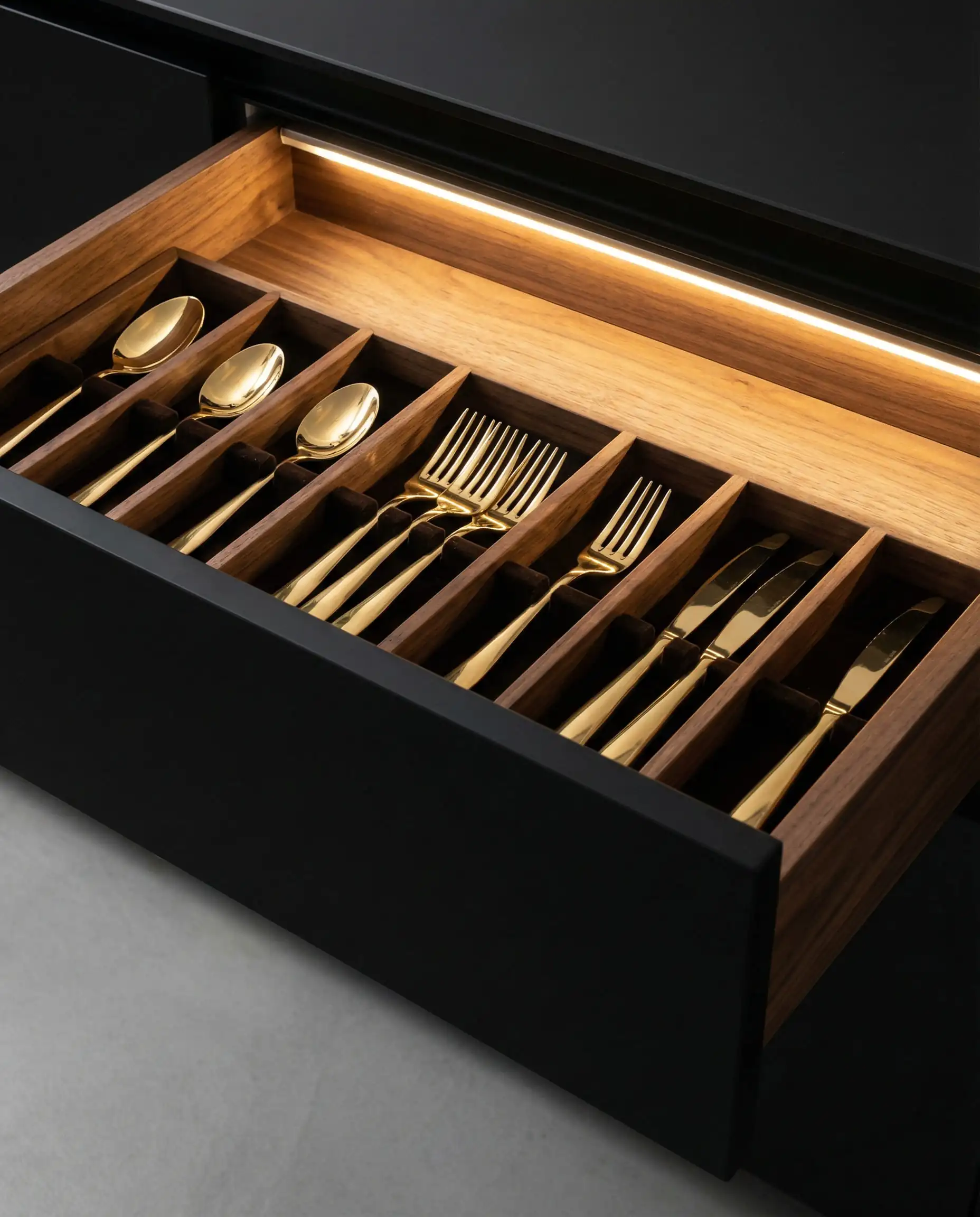 Open kitchen drawer with walnut interior and LED lighting illuminating gold cutlery.