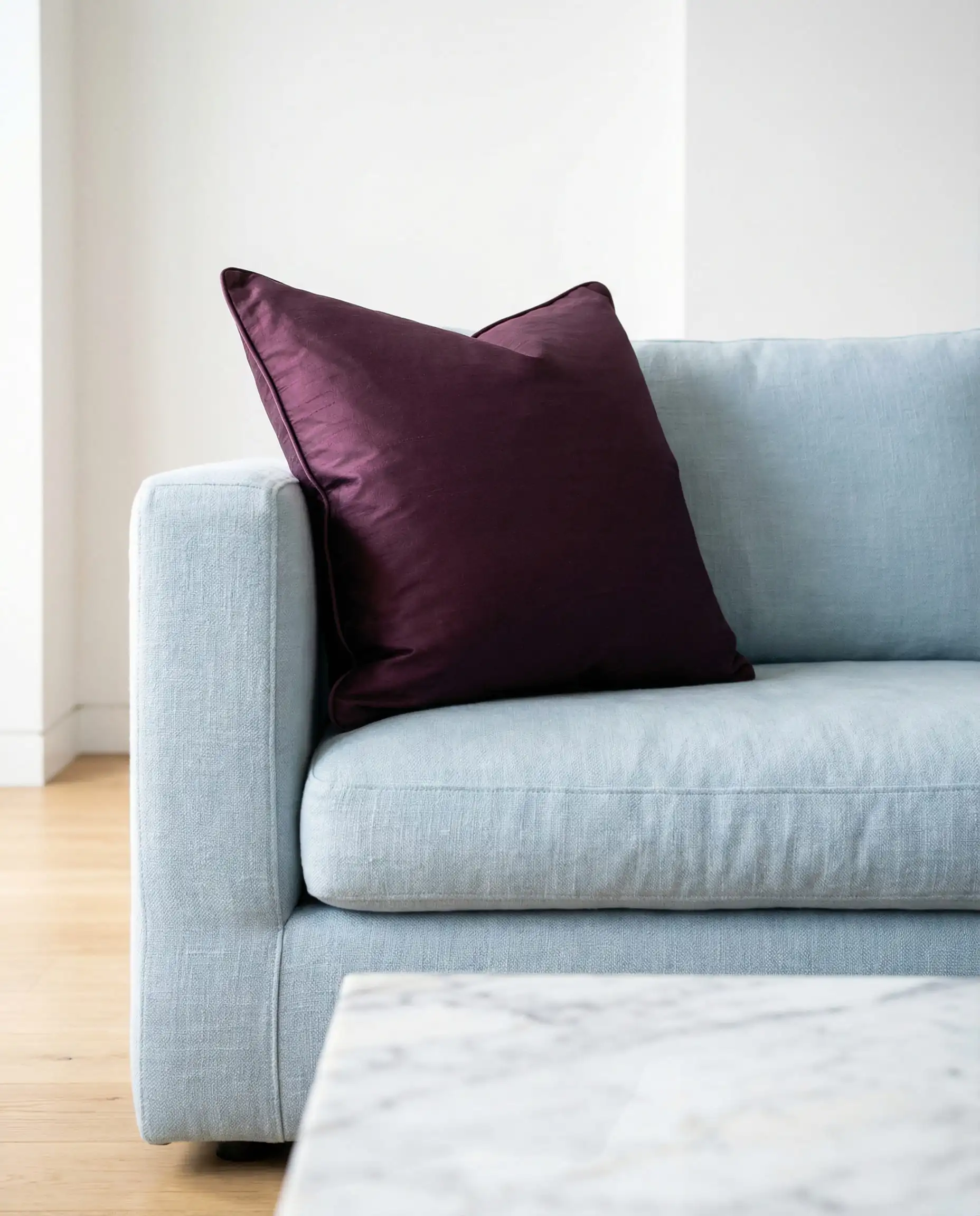 A pastel light blue linen sofa contrasting with a deep plum purple silk pillow in a modern living room.