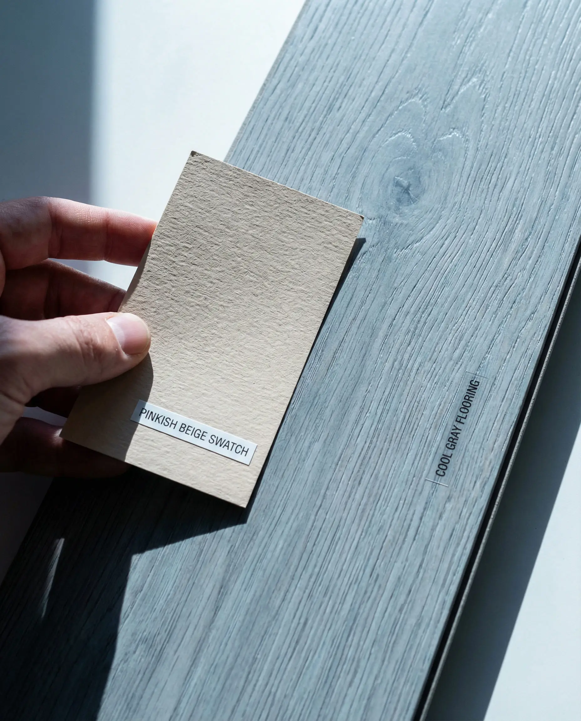 Close up of a pinkish-beige paint swatch clashing visibly against a cool blue-toned gray floor plank.