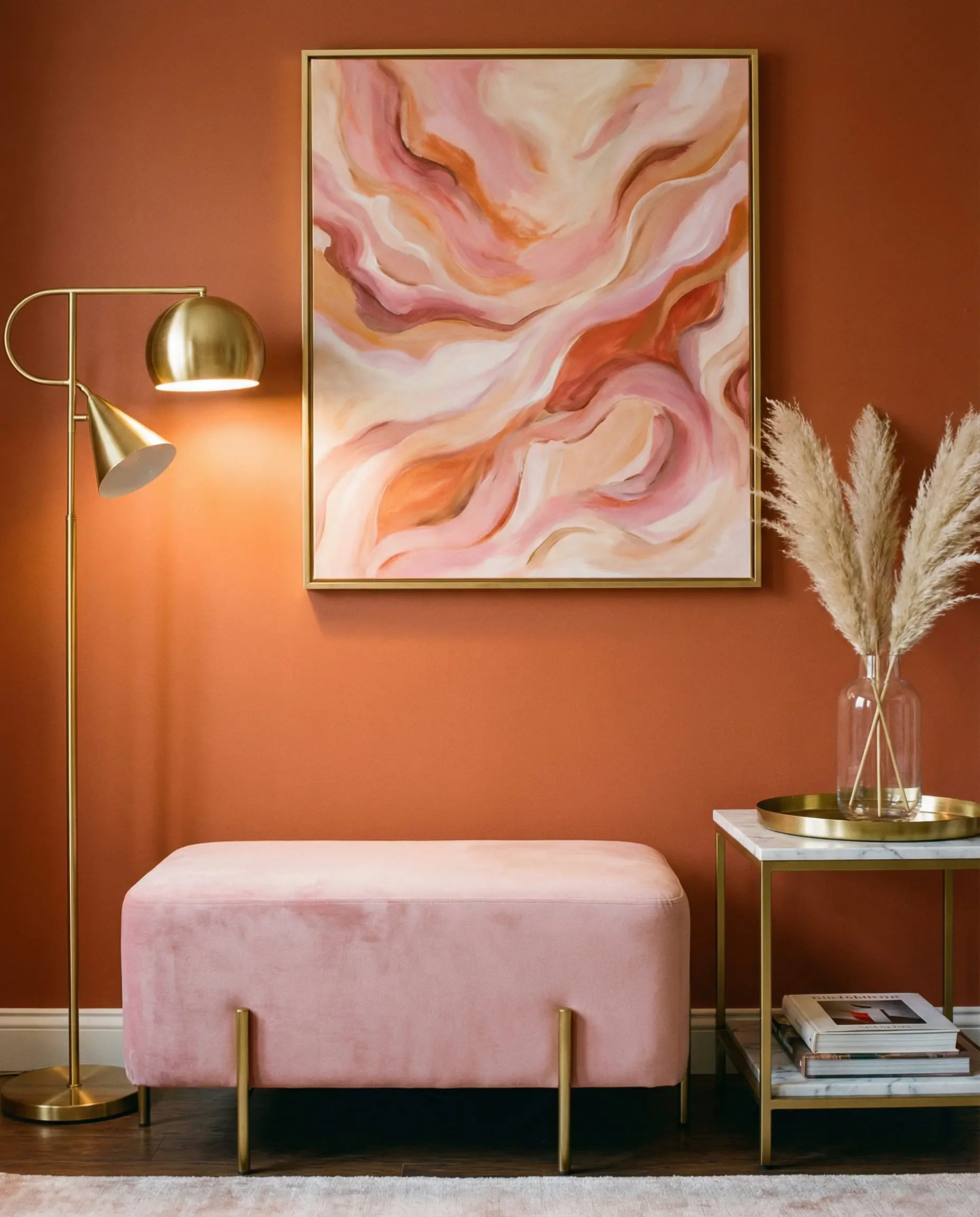 Chic living room vignette with a soft pink velvet ottoman and gold accents against a rust orange wall.