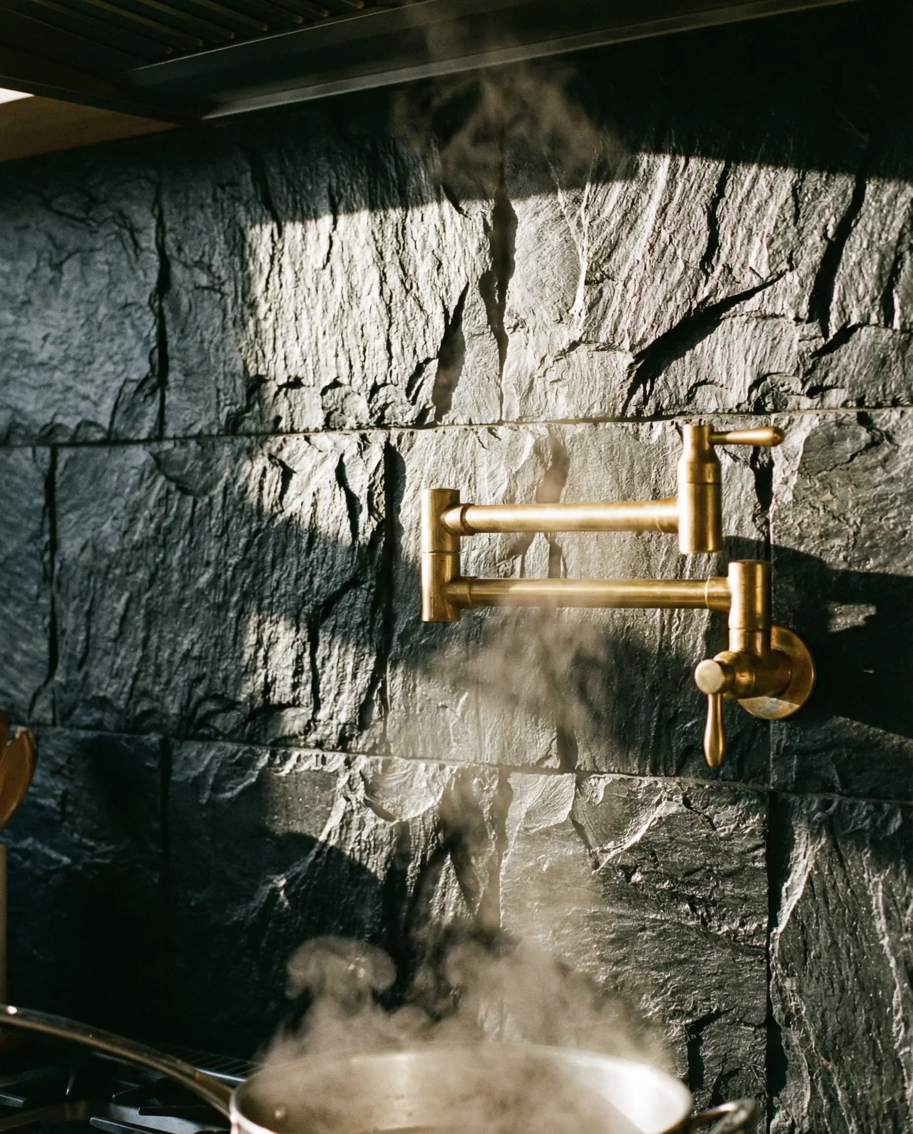 Rough-hewn black slate tile backsplash with a shiny unlacquered brass pot filler faucet.
