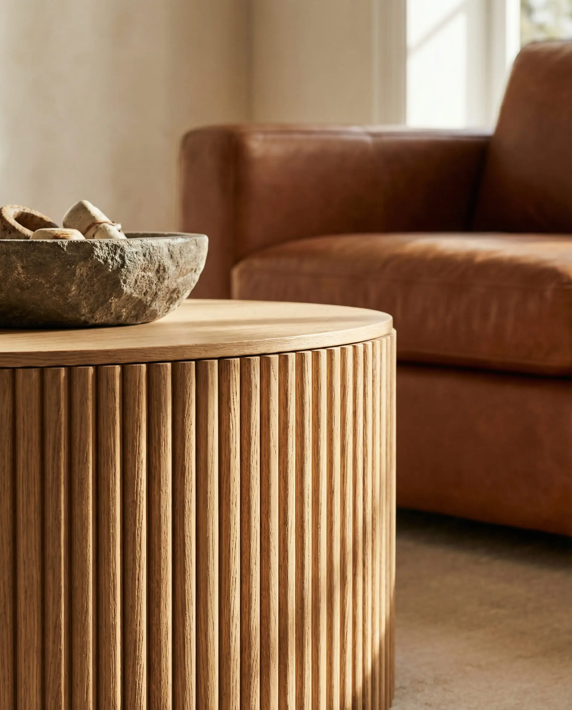 Close up texture shot of a round coffee table with a vertical slatted white oak base next to a cognac leather sofa.